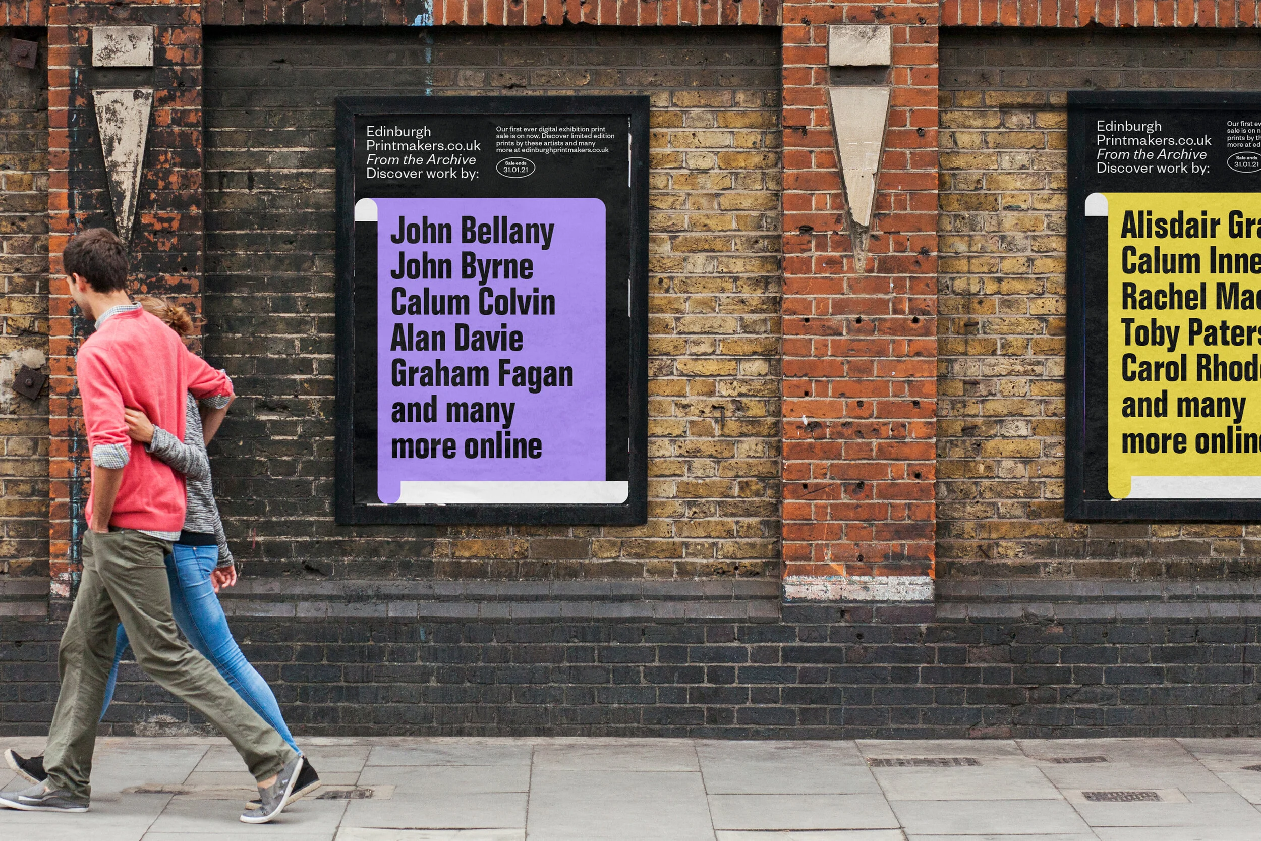 Two people walking past brick walls with framed posters listing names of artists and online contributors.