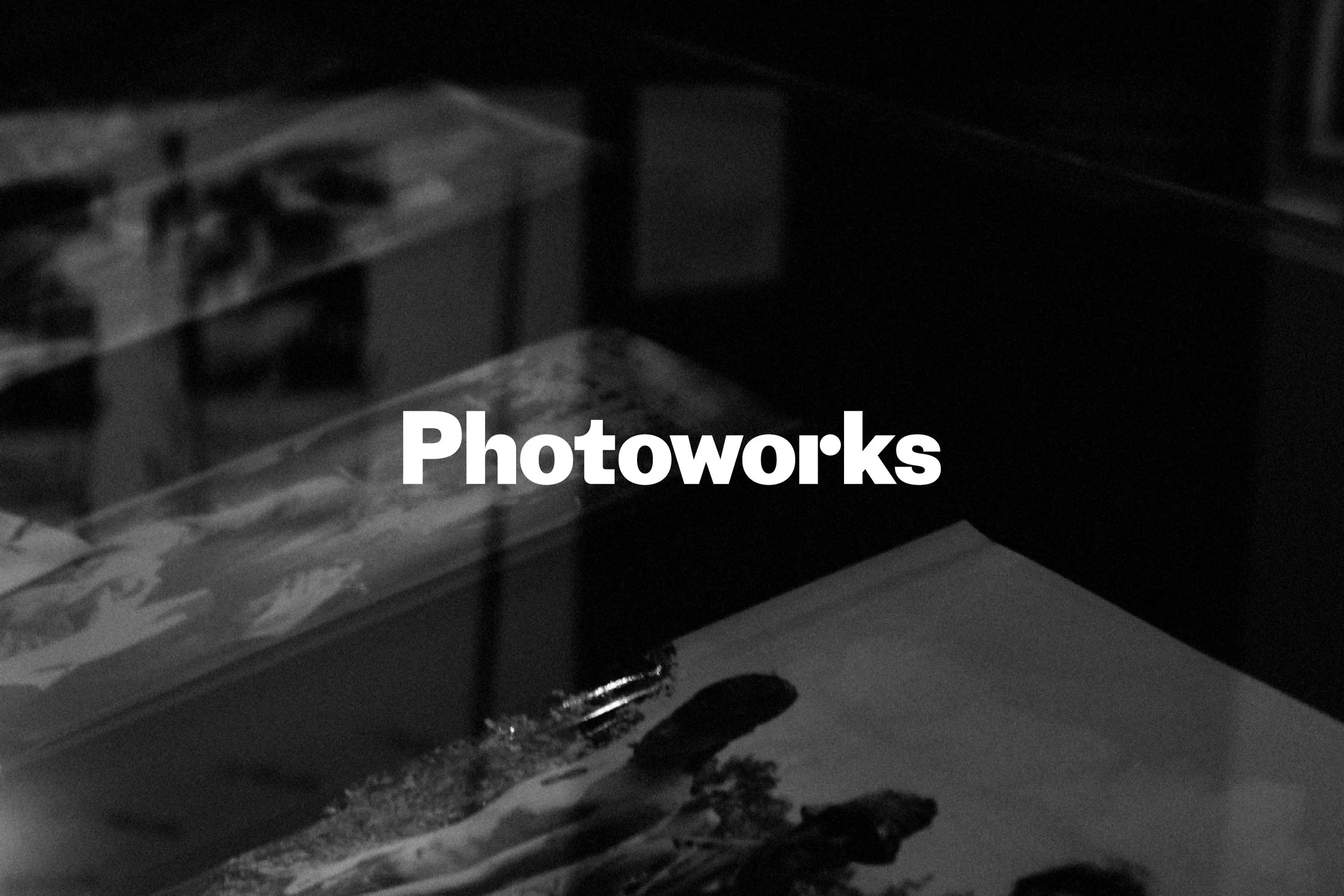 Black and white photo of a desk with a book, a keyboard, and a glass of water, with the word 'Photoworks' in bold white letters across the center.
