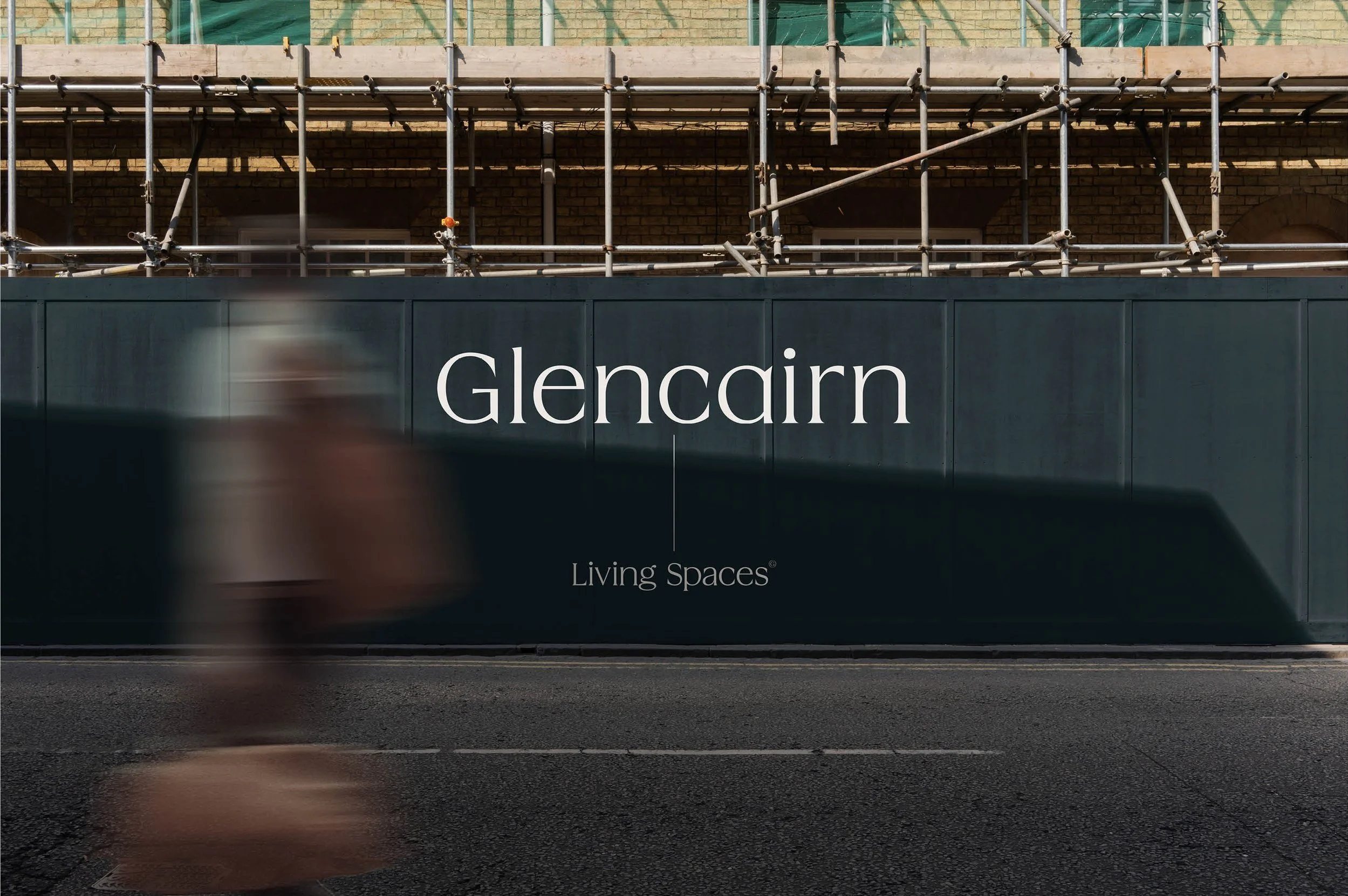 A blurred person walking past a dark green barrier with the word 'Glencairn' and under it, the phrase 'Living Spaces' on it. Behind the barrier, there is a building under construction with scaffolding.
