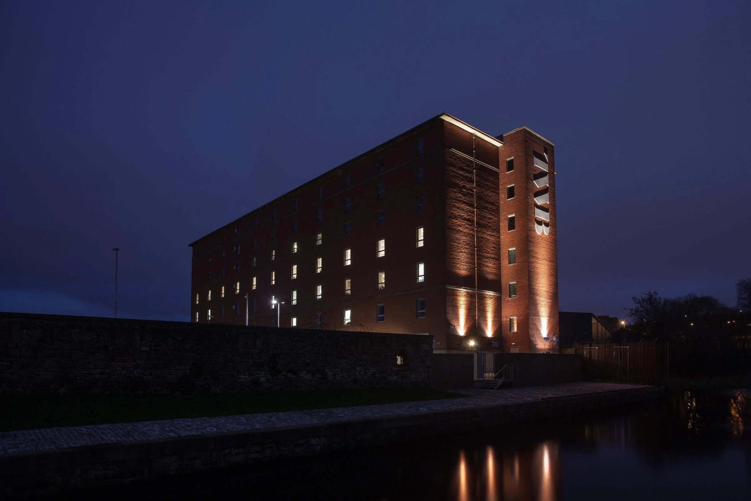 Nighttime view of a tall brick building illuminated by exterior lights, with several windows lit up, adjacent to a canal reflecting the scene.