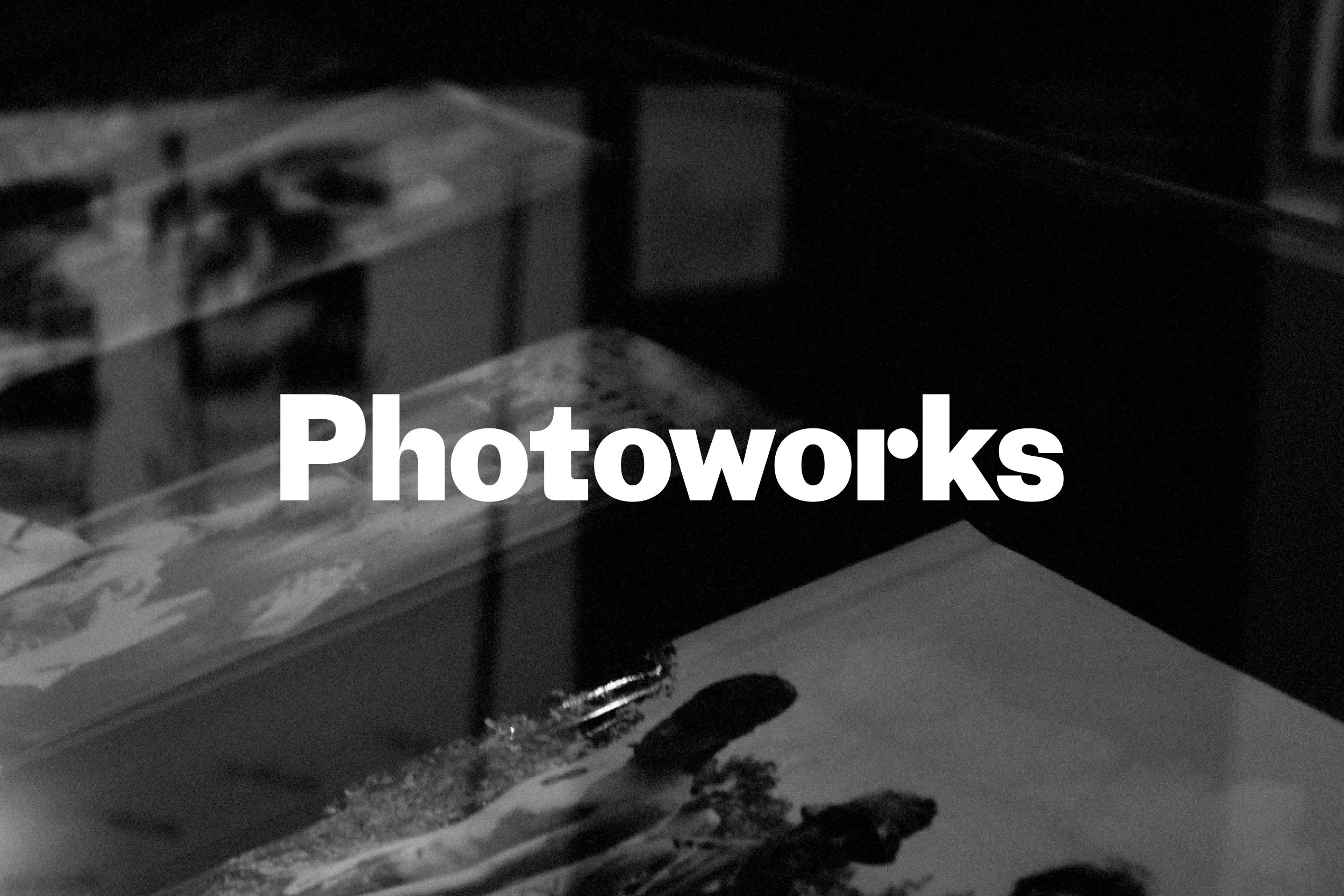Black and white photo of a desk with a magazine, a photo print of a person, and a pen, labeled 'Photowork's' in white text.