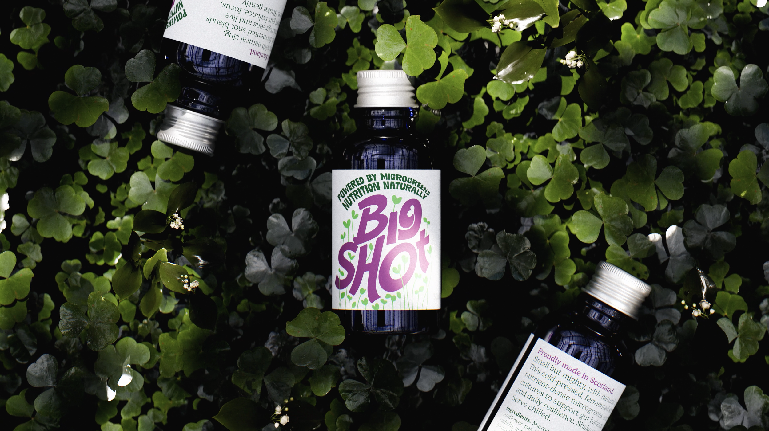 Three small dark bottles of BIO SHOT nutrient drink lying on green clover leaves outdoors.