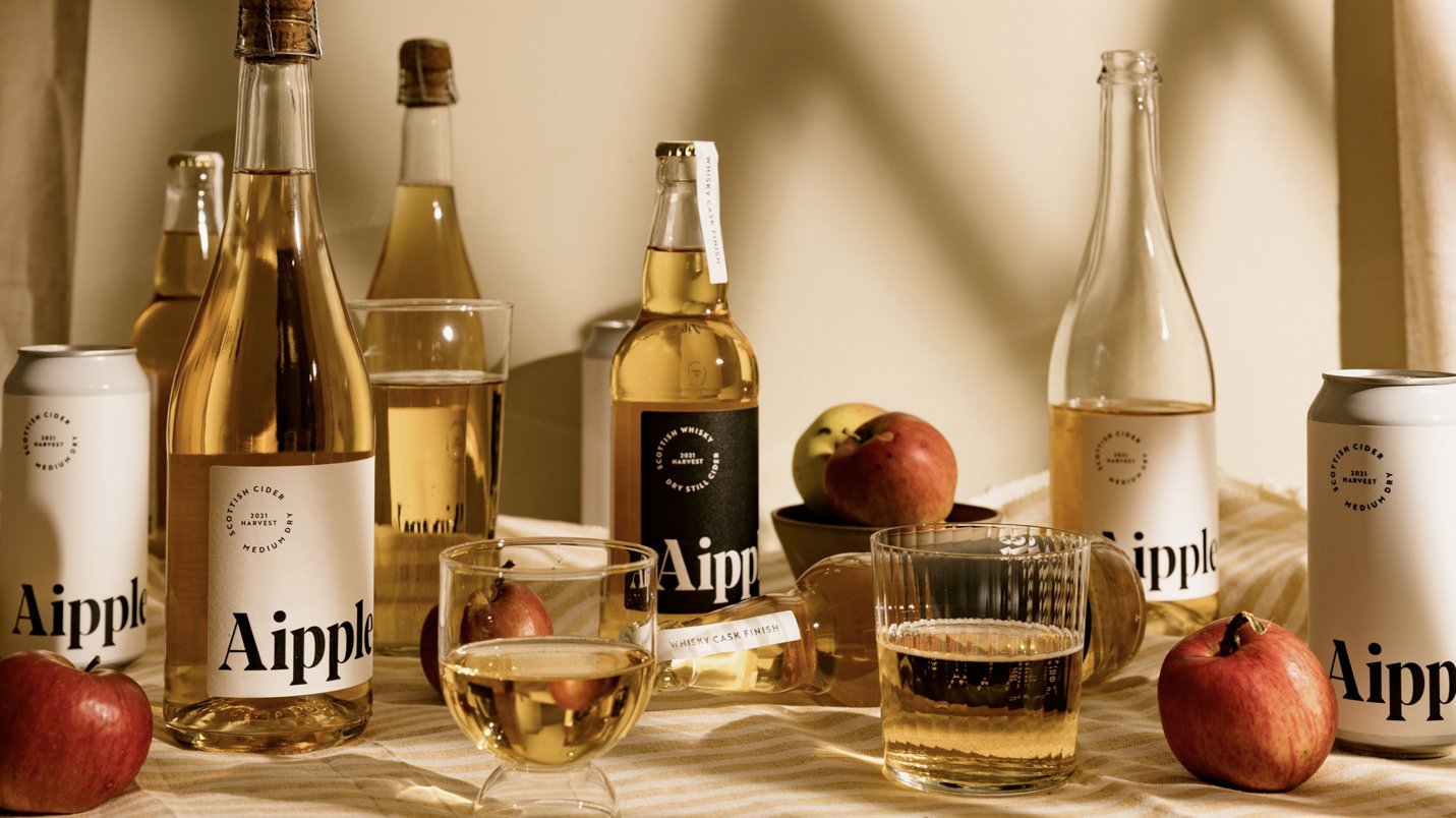 Arrangement of bottles, cans, and glasses of apple cider, along with apples and a bowl of fruit on a table.