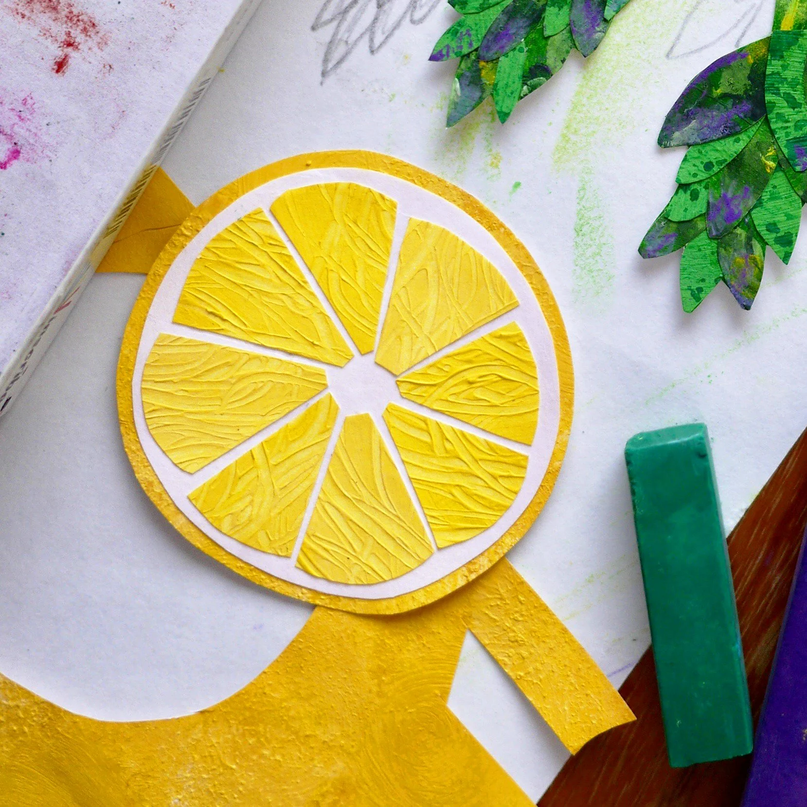 A craft project featuring a paper lemon slice with yellow textured sections and white borders, surrounded by colorful paper leaves and craft supplies.