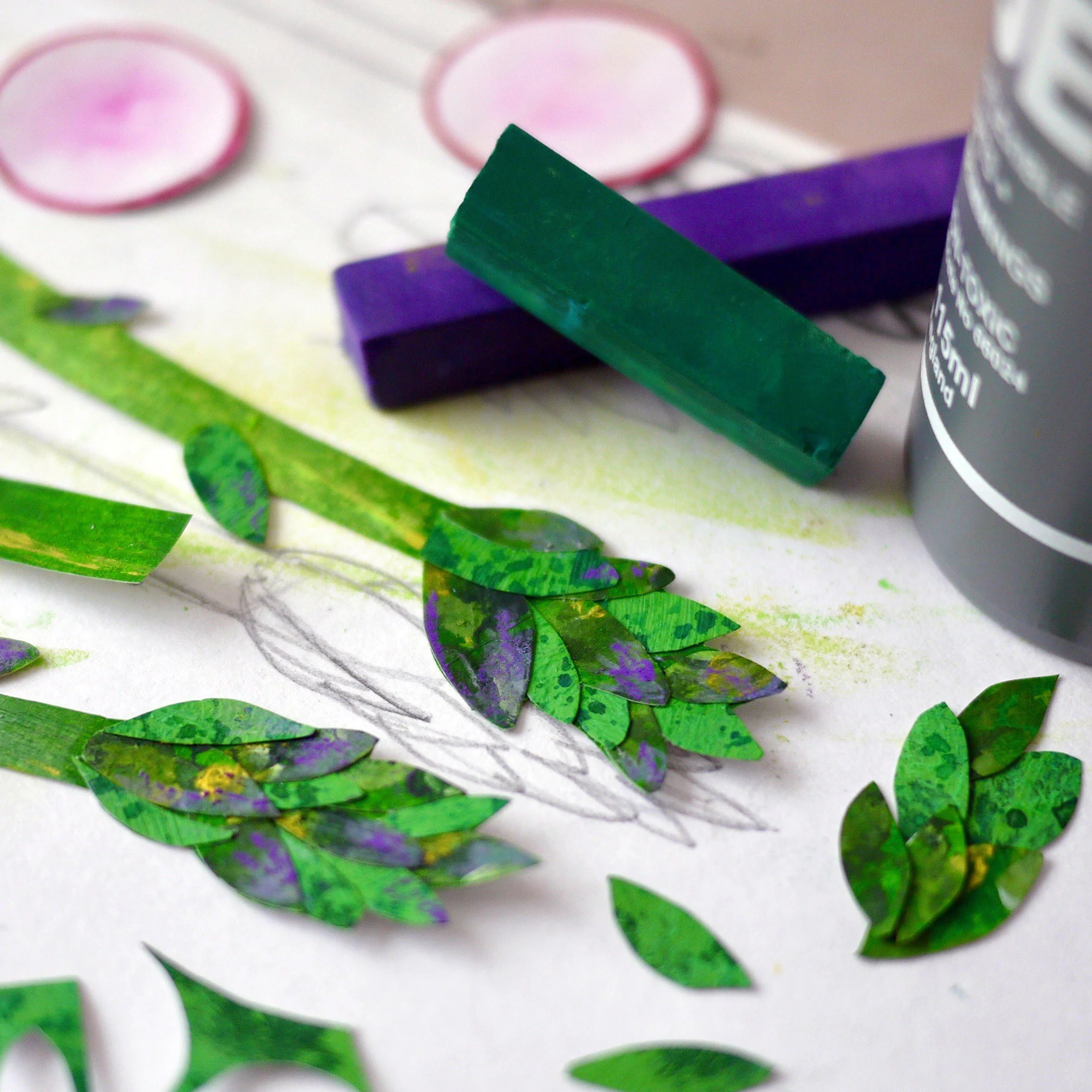Green, purple, and black paper cutouts resembling leaves, a green and purple chalk stick, a partially visible bottle, and a faint sketch of a plant on white paper.