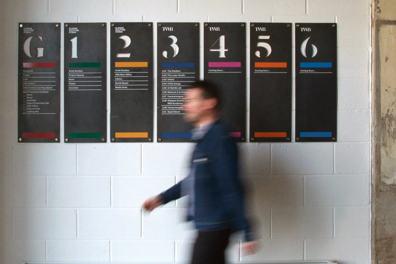 A person walking past a wall with black directory signs at Glasgow Sculpture Studios, with room numbers G1, 2, 3, 4, 5, 6, and accompanying text indicating various studio names and functions.