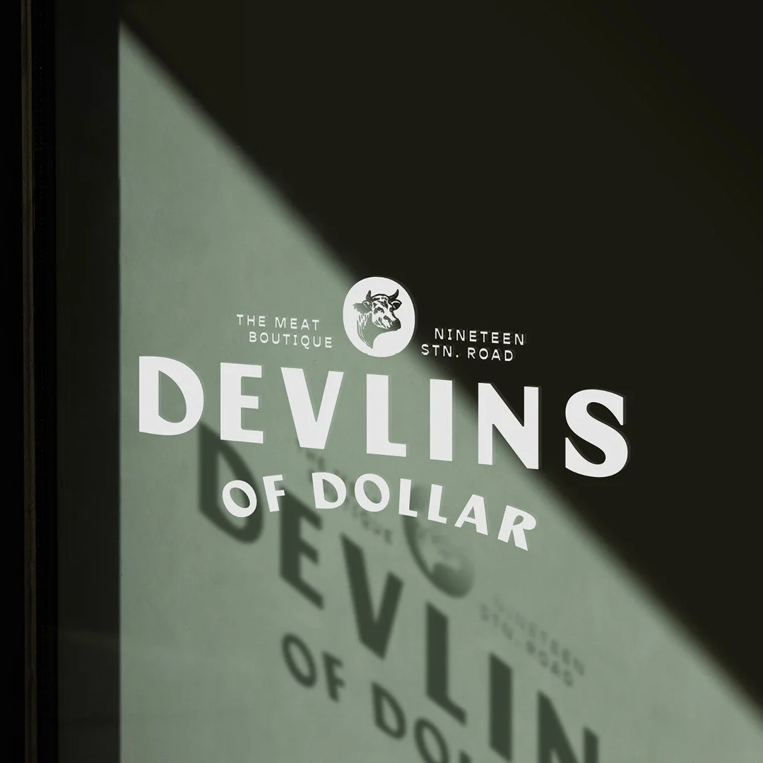 Close-up of a sign with the text 'Devlins of Dollar,' a logo of a wolf or dog, and details about a business called The Meat Boutique, located at Nineteen Stn. Road.