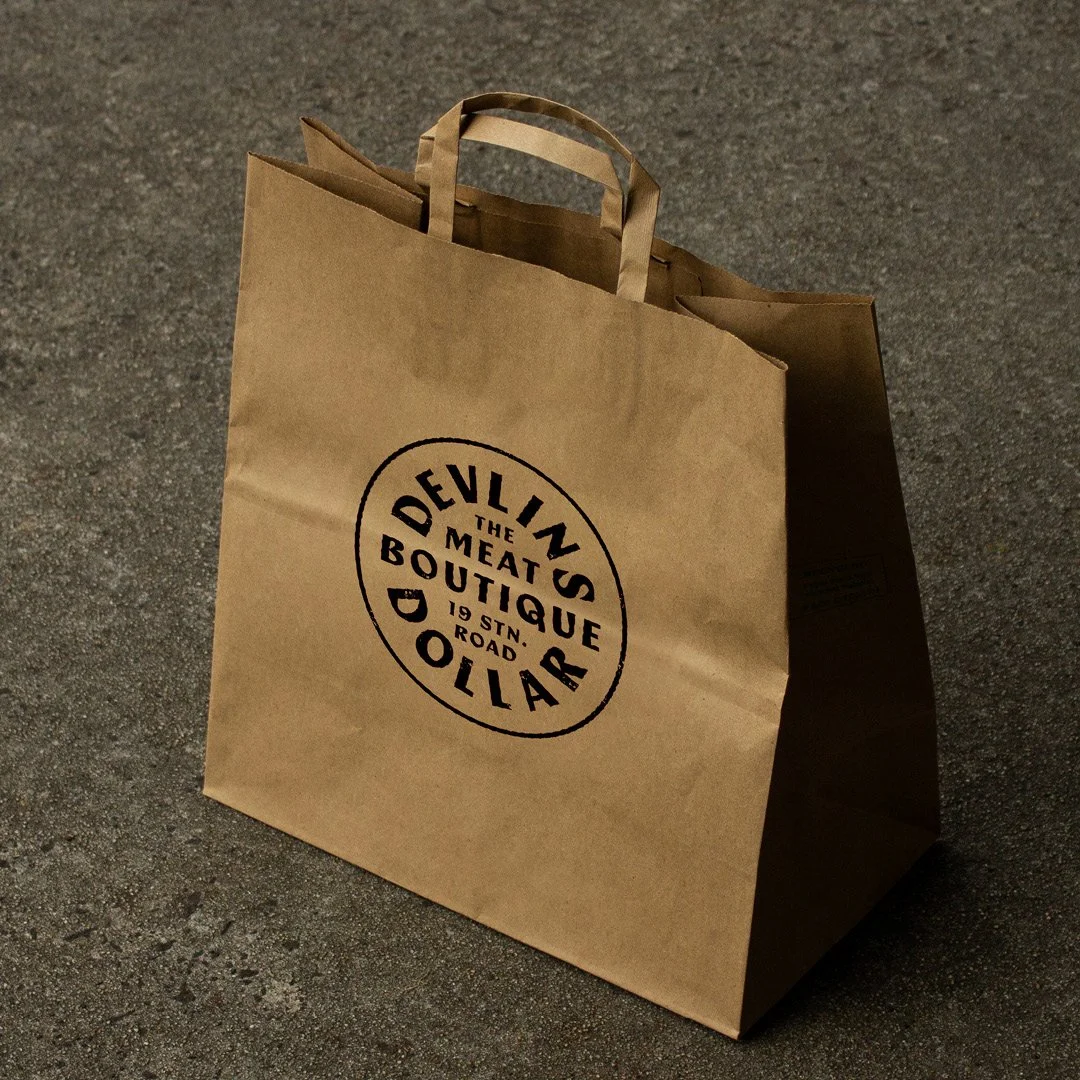 Brown paper shopping bag with black logo reading 'The Meat Boutique, 19 Stn Road, DolLAR, Devin's' on a rough gray ground.