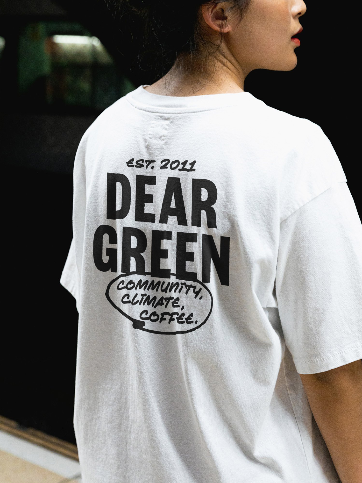 A person wearing a white T-shirt with black text that reads 'Dear Green' with a speech bubble that says 'Community, Climate, Coffee.' The person has dark hair and is shown in profile against a dark background.