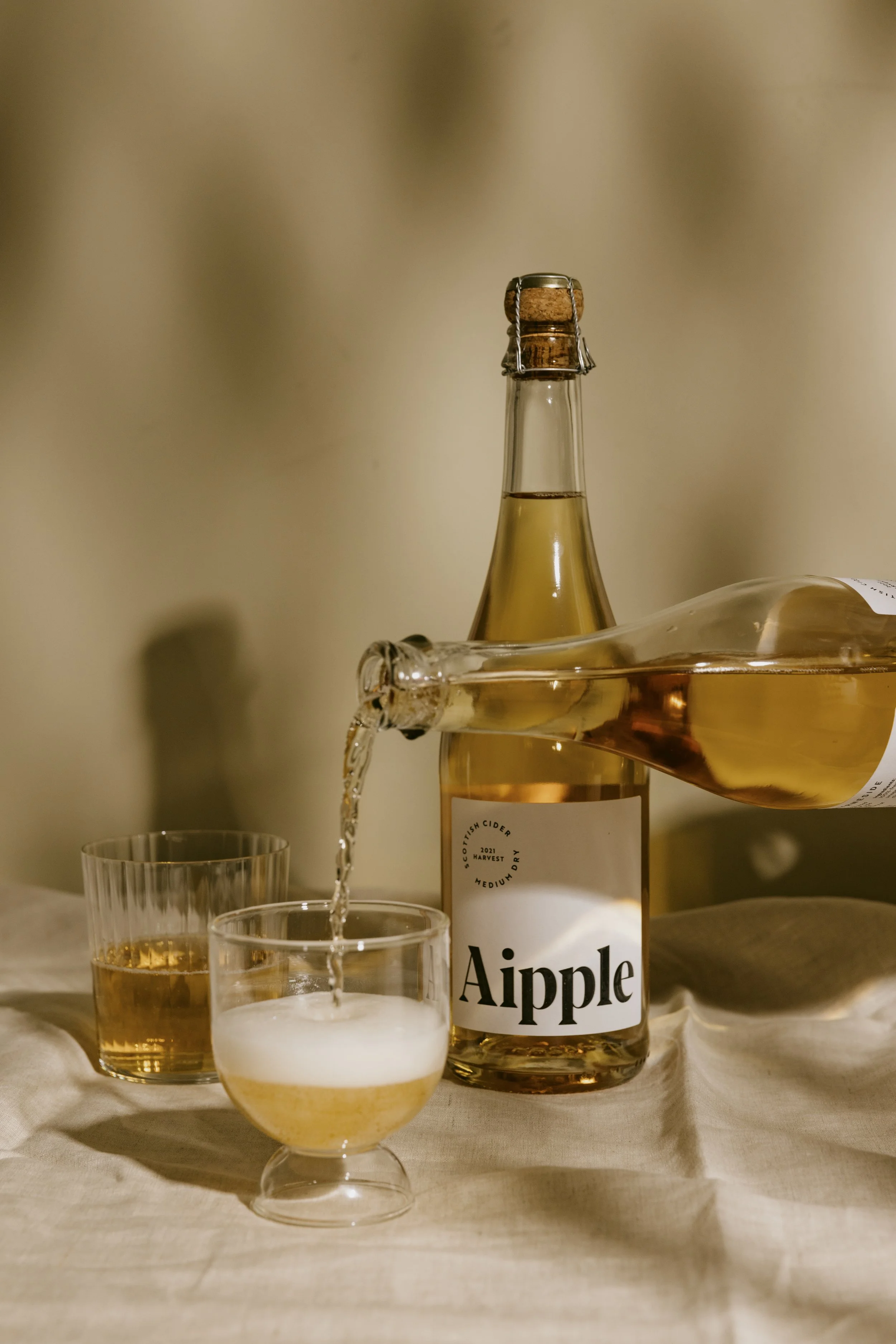 A bottle labeled "Aipple" with light-colored cider, two glasses filled with cider, one of which is being poured, on a neutral tablecloth background.