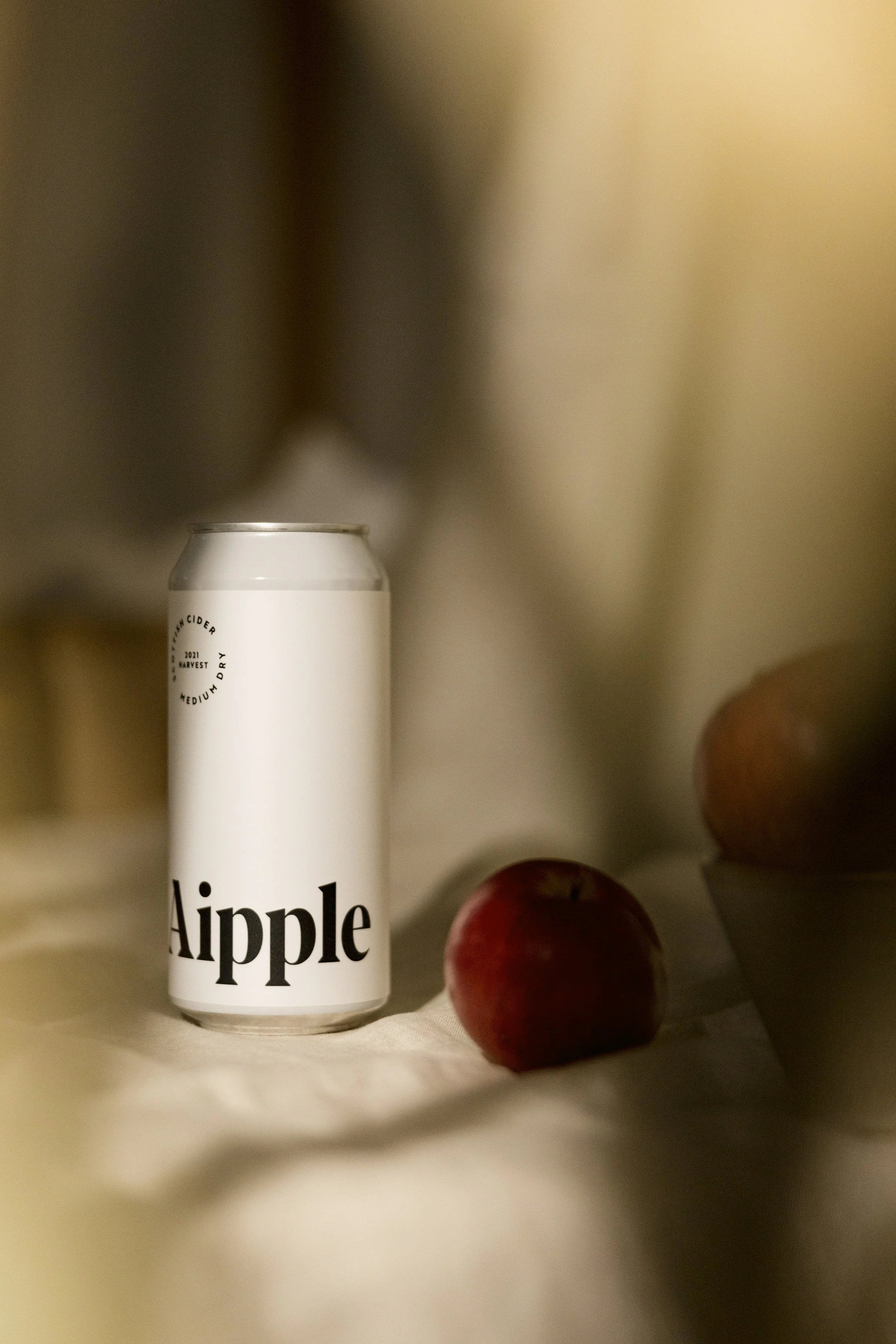A can of apple-flavored beverage with minimalist white label and black text, placed next to a red apple on a white cloth surface, with blurred background.