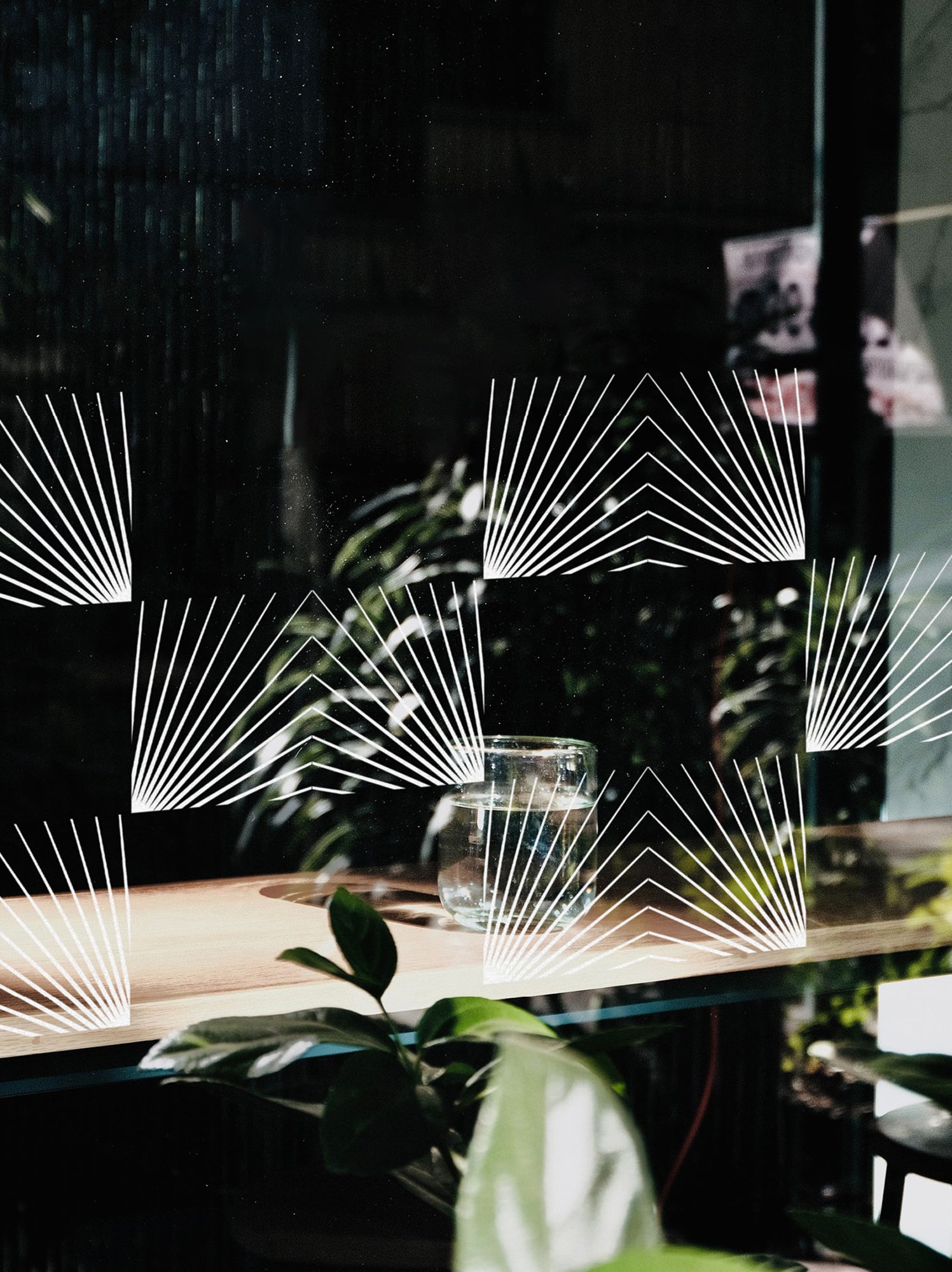 A glass of water on a wooden surface with decorative white geometric lines, plants, and reflections in the background and glass.