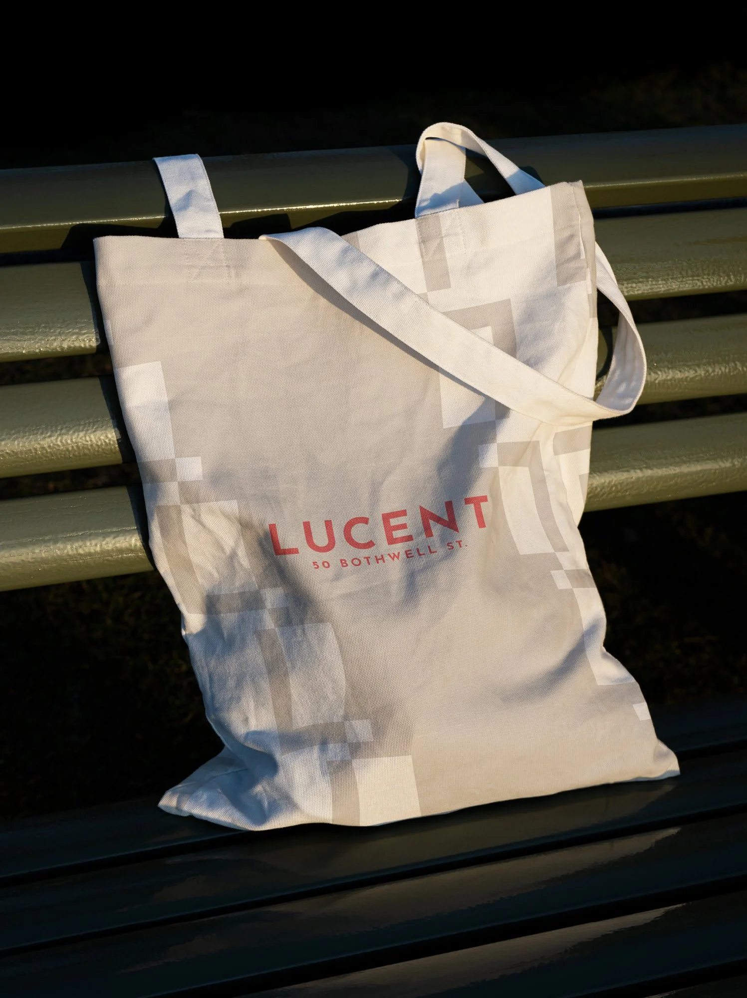 A white tote bag with the word 'LUCENT' and address '50 Bothwell St.' printed in red, hanging on a green park bench.