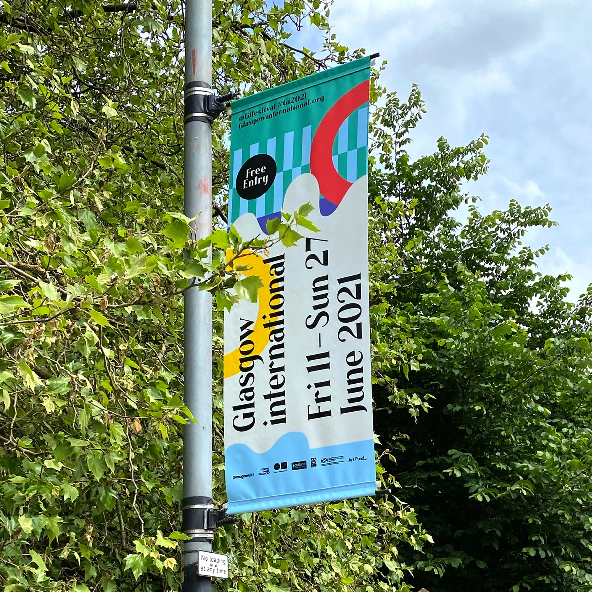 Festival banner promoting Glasgow International 2021, taking place from June 11 to June 27, with free entry, attached to a street pole surrounded by green tree leaves.