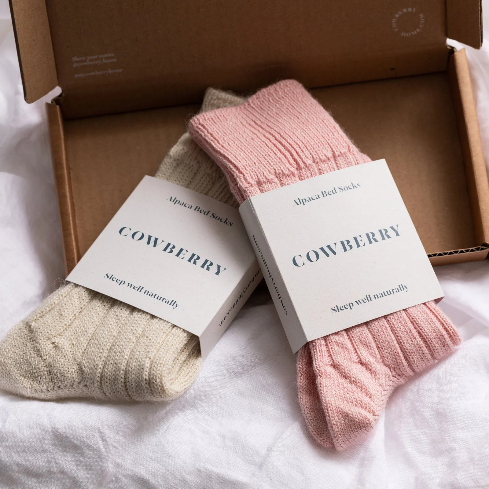 Two pairs of alpaca bed socks, one beige and one pink, with labels reading 'Cowberry' and 'Sleep well naturally,' in a brown cardboard box on a white surface.