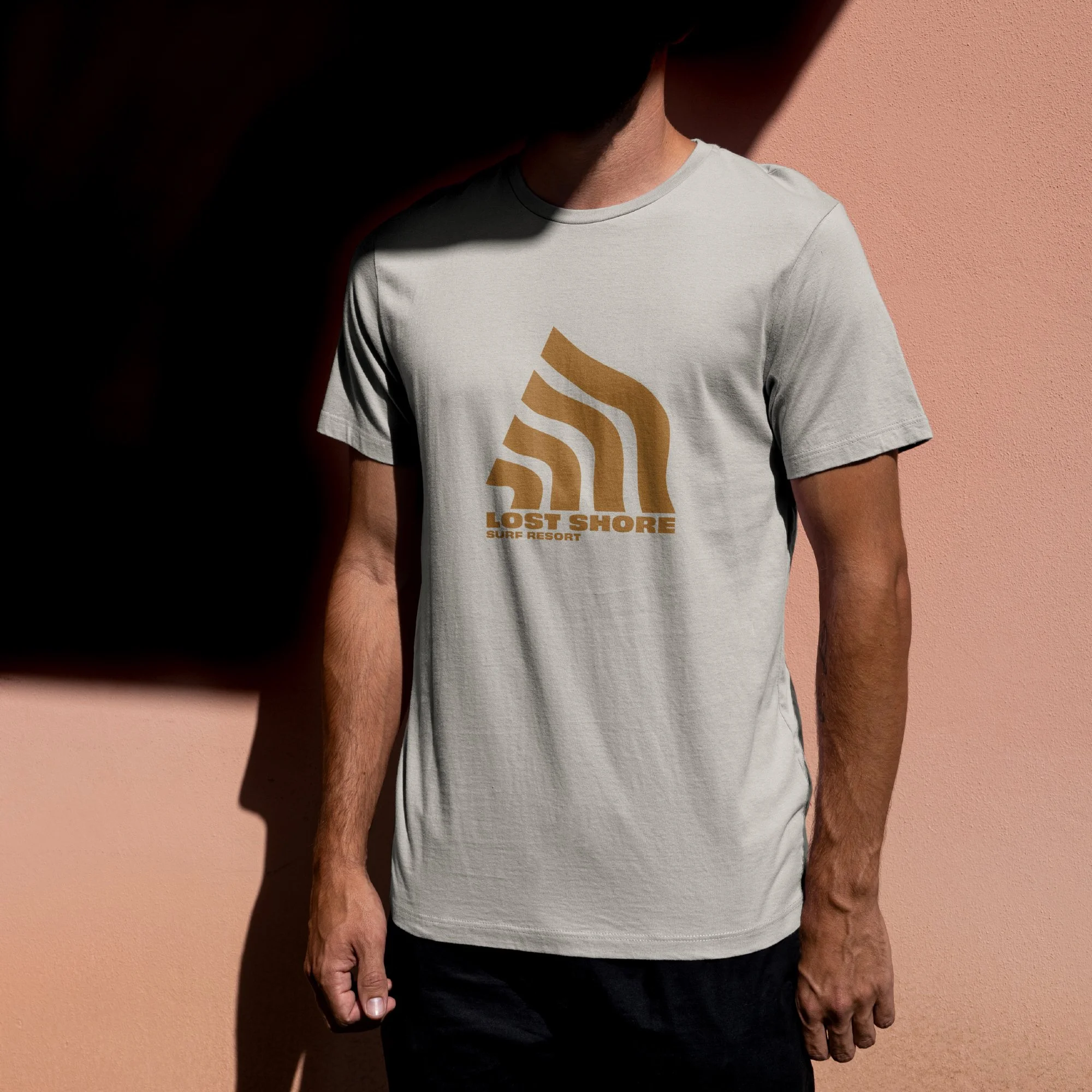 Person wearing a light-colored T-shirt with 'Lost Shore Surf Resort' logo on the front, standing against a pink wall with shadow.