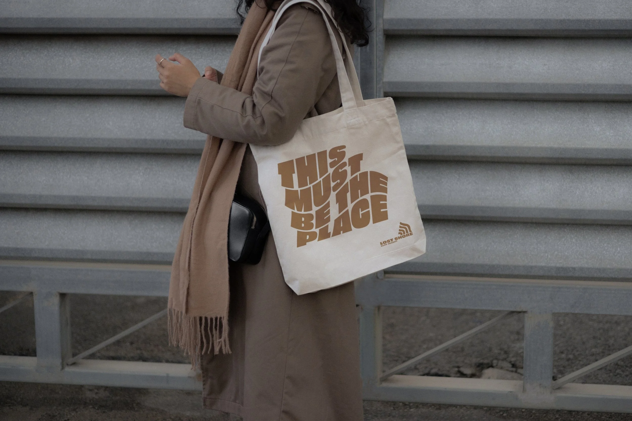 Person standing outdoors, carrying a beige tote bag with the words 'This Must Be The Place' printed on it, dressed in a beige coat with a light brown scarf.