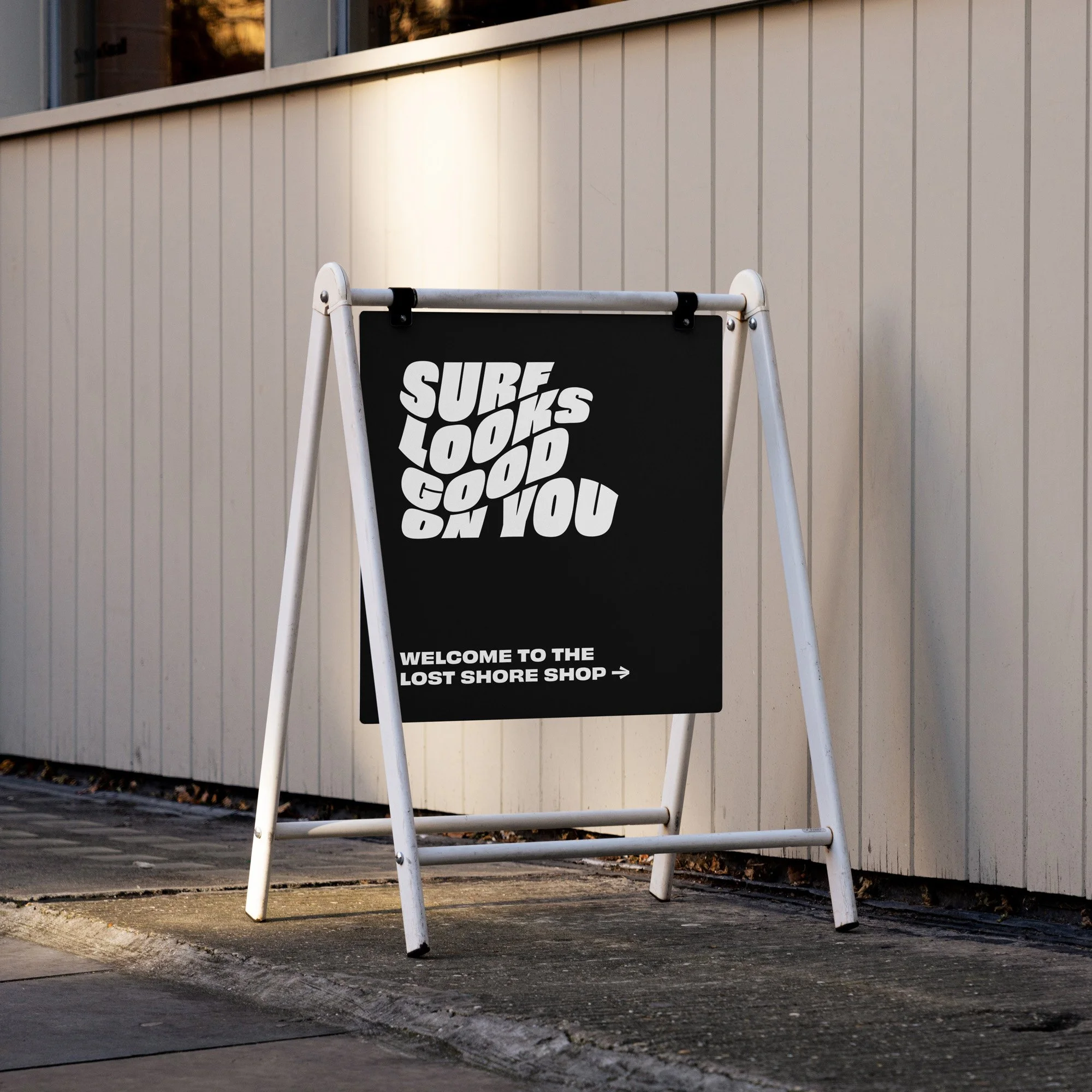 A black sandwich board sign on a sidewalk promoting the Lost Shore Shop with the phrase 'Surf Looks Good on You' in bold white letters.
