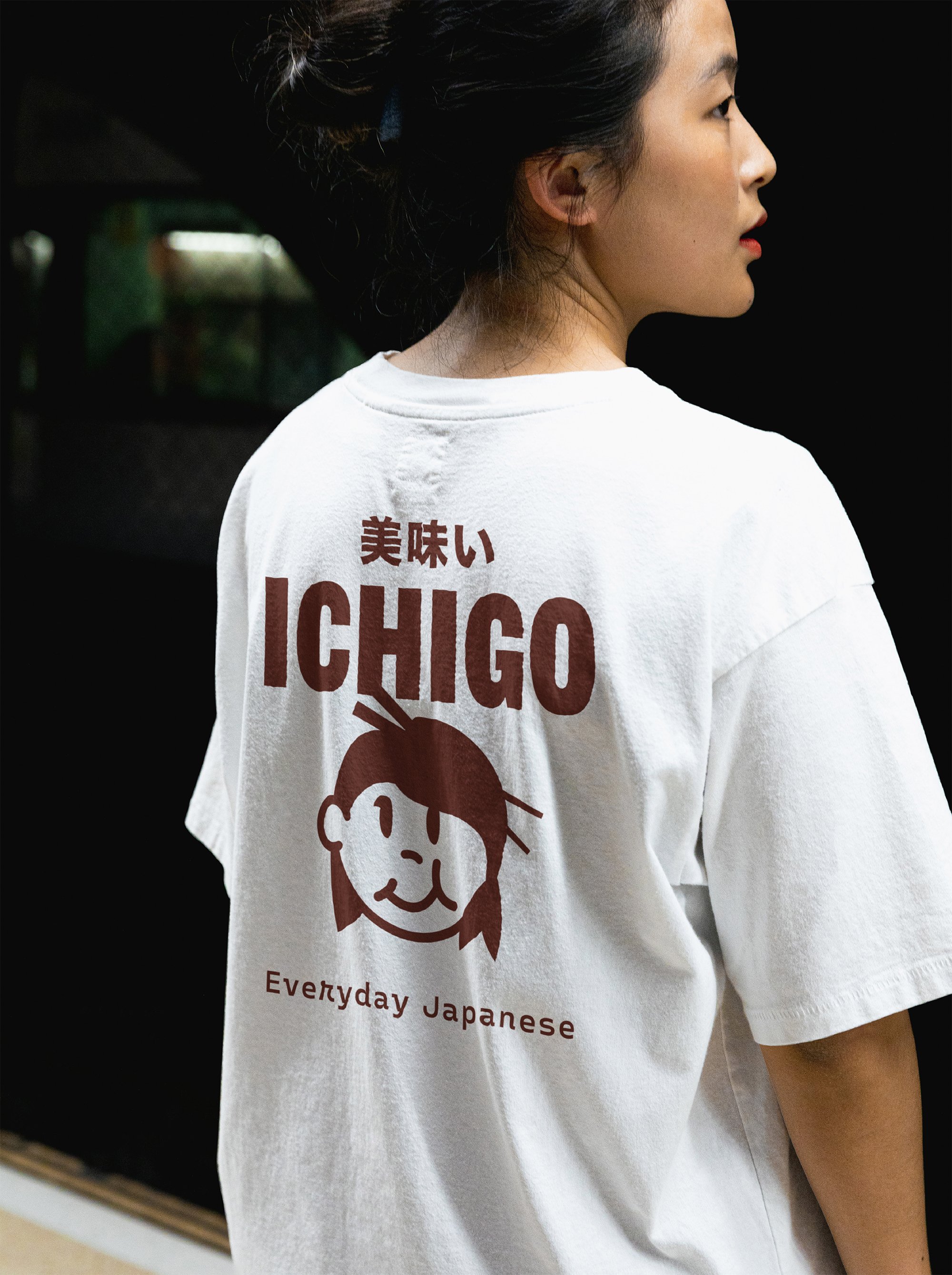 A woman wearing a white T-shirt with Japanese characters, a cartoon girl's face, and the words 'ICHIGO' and 'Everyday Japanese' printed on the back.