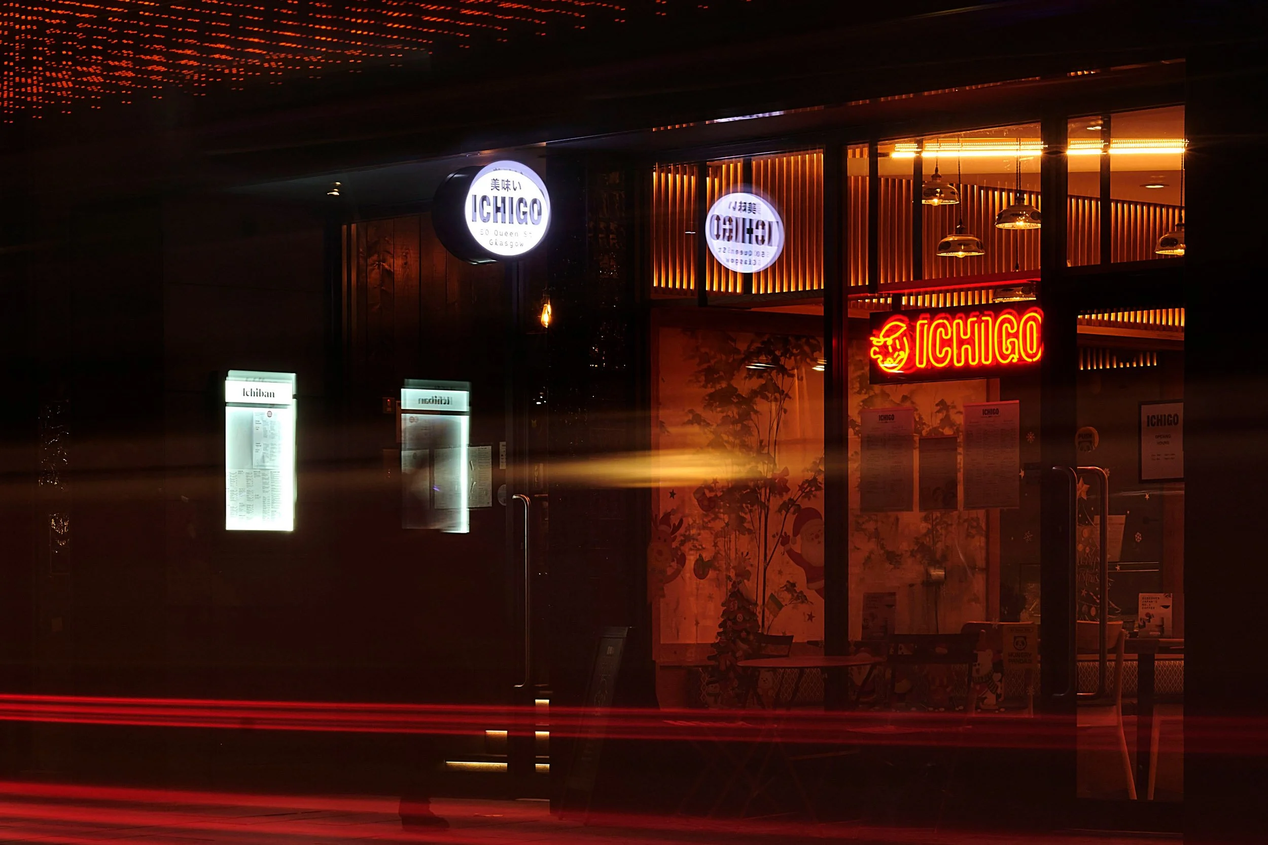 Nighttime view of a restaurant called Ichigo, with illuminated signs and menu boards visible through the glass facade, and light trails from passing vehicles outside.