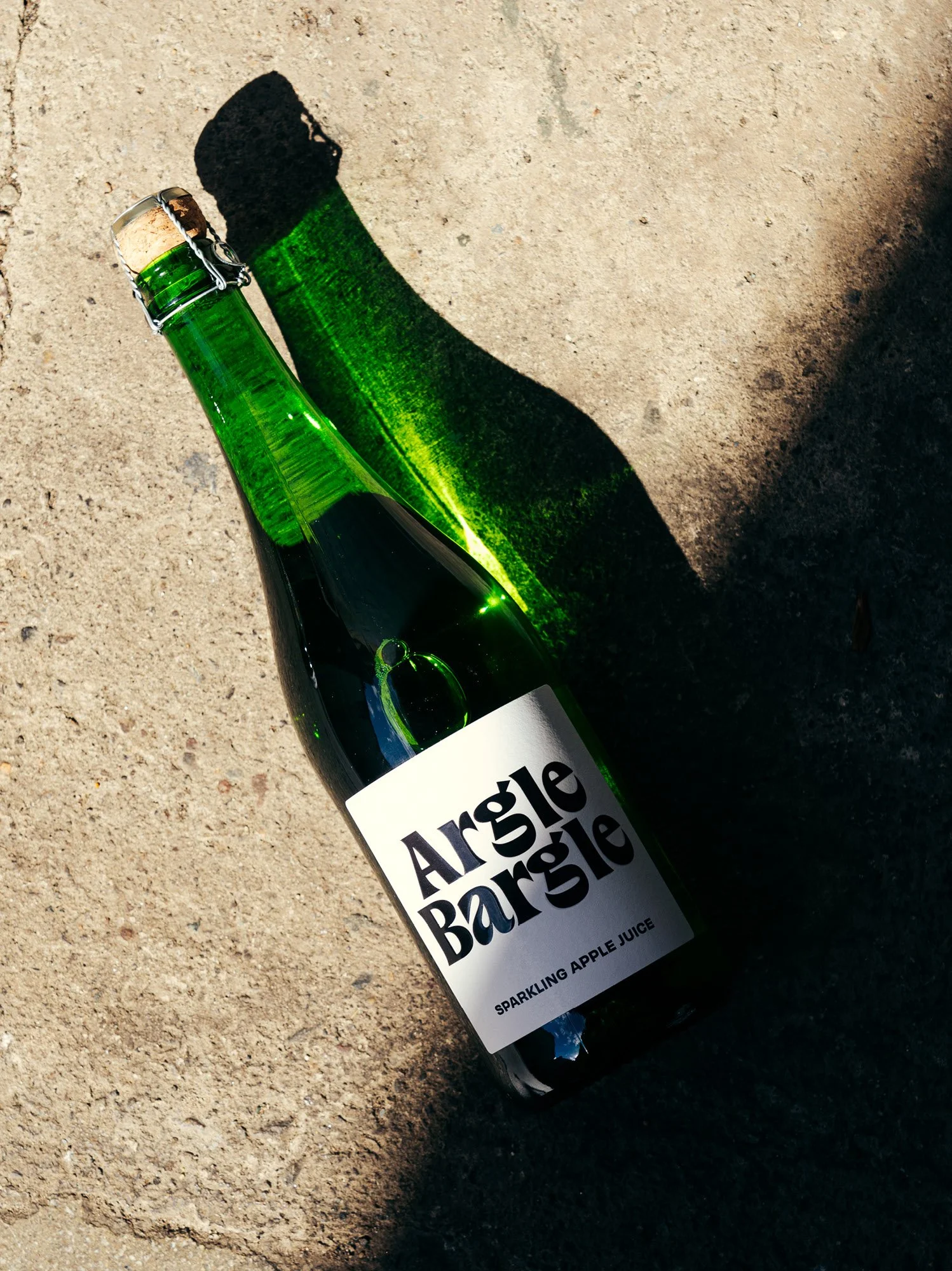 A green glass bottle of Argle Bargle sparkling apple juice lying on a sandy surface, casting a shadow.