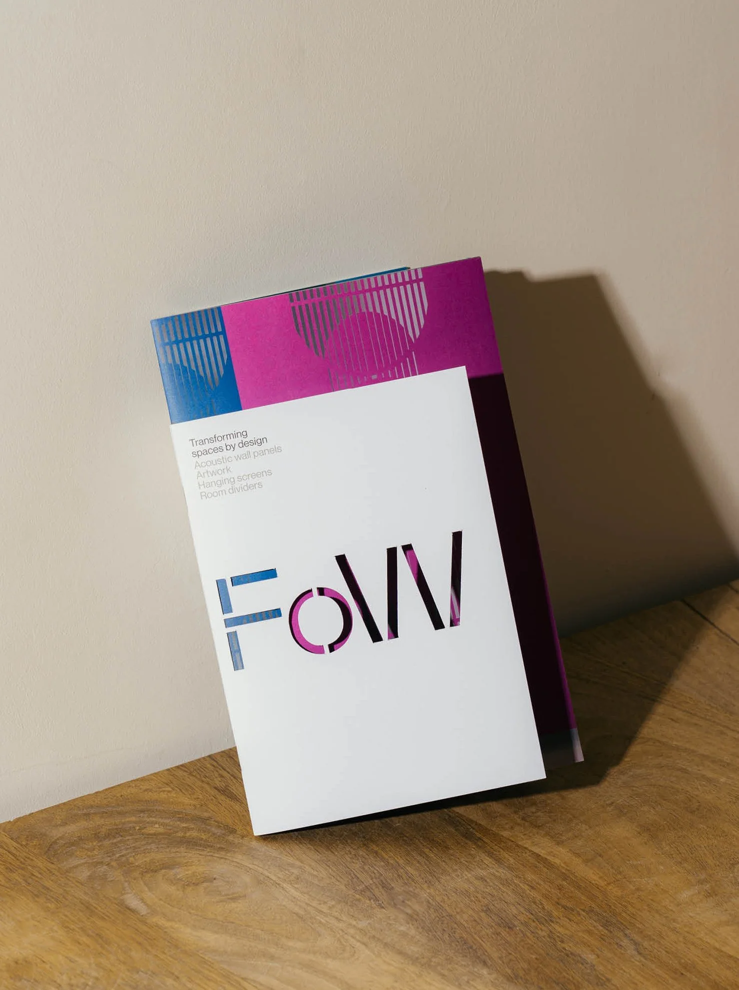 Brochure titled 'FORM' with abstract design in pink, purple, black, white, and blue colors, leaning against a beige wall on a wooden surface.