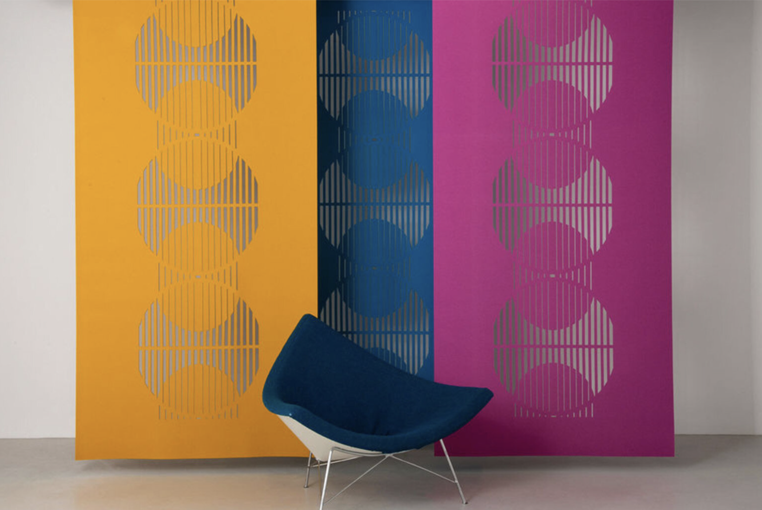 Colorful wall with three panels in yellow, blue, and pink, featuring geometric cutouts of circles and lines, with a modern blue chair in front.