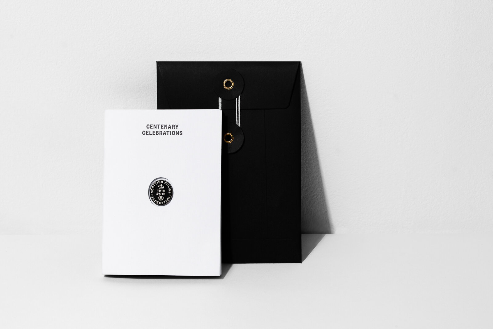 A white booklet titled "Centenary Celebrations" and a black envelope with gold accents on a white surface and wall.