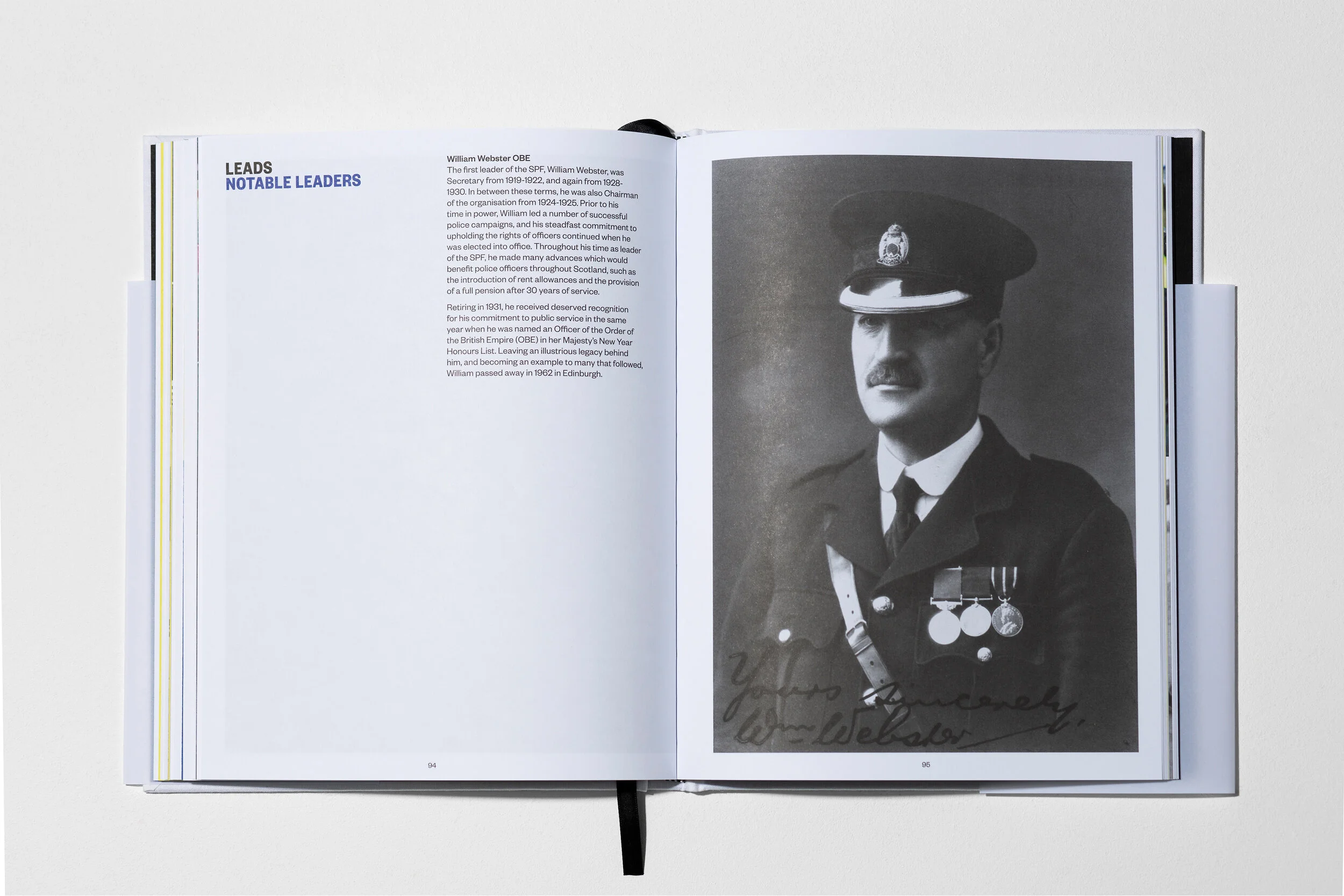 An open book showing a black and white photograph of a man in a police uniform with medals and a cap, accompanied by a signature on the photo, and pages with text about William Webster.