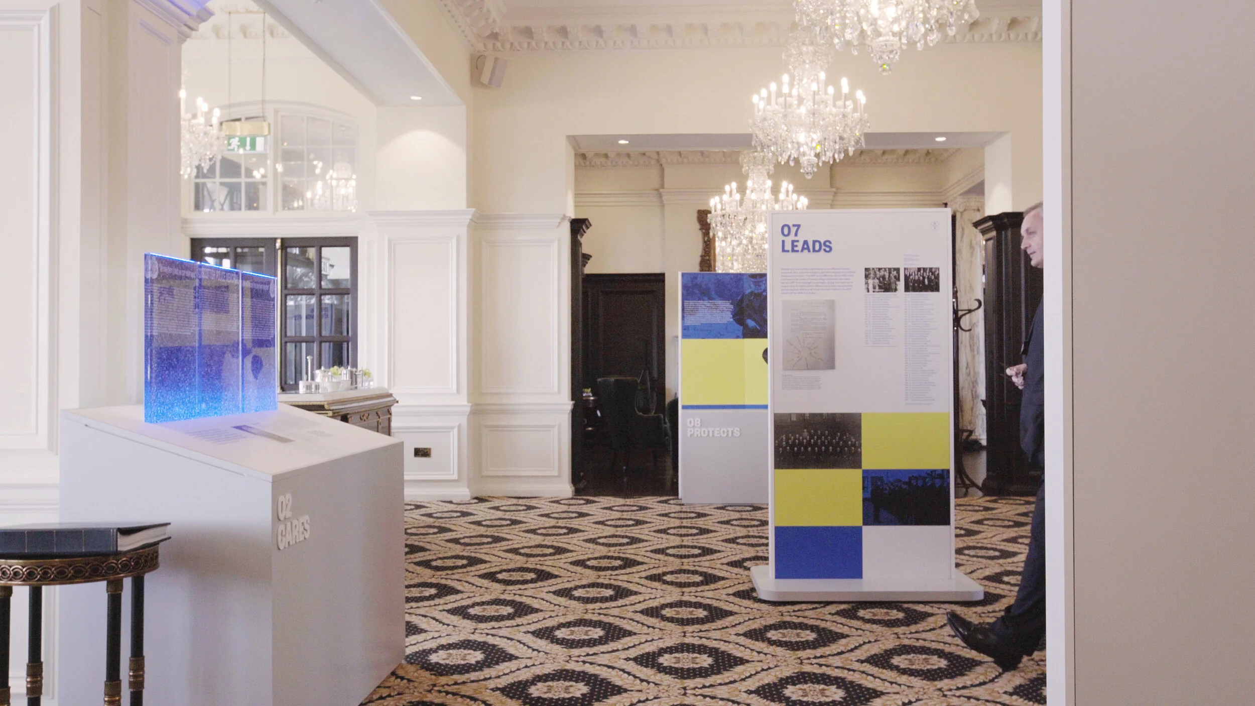 Art exhibition space with informational display boards, chandeliers, ornate ceiling molding, and patterned carpet, with a person partially visible on the right side.