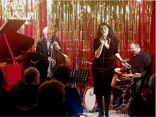 Junge weibliche Sängerin singt auf einer Bühne mit drei Musikern, die in jazziger Atmosphäre spielen, vor goldener Dekorationshintergrund.