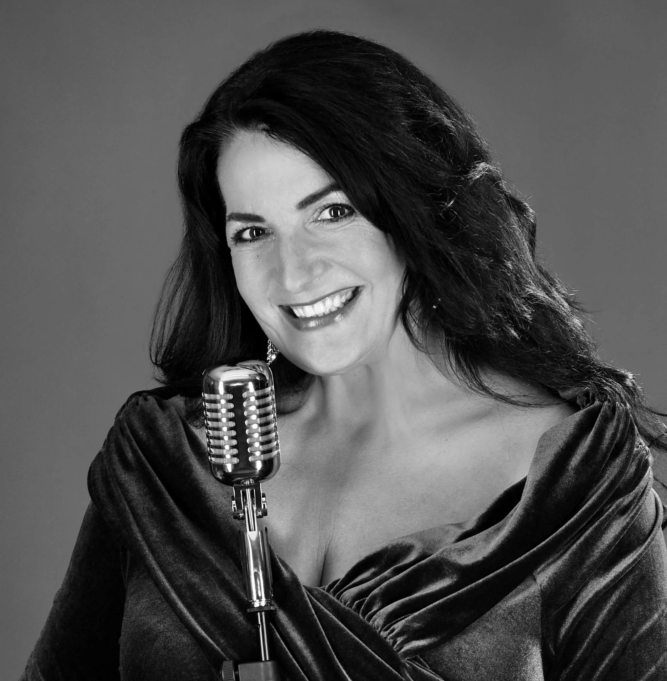 Schwarze und weiße Aufnahme einer lachenden Frau mit dunklem, lockigem Haar, in eleganter Kleidung, vor einem Mikrofon.