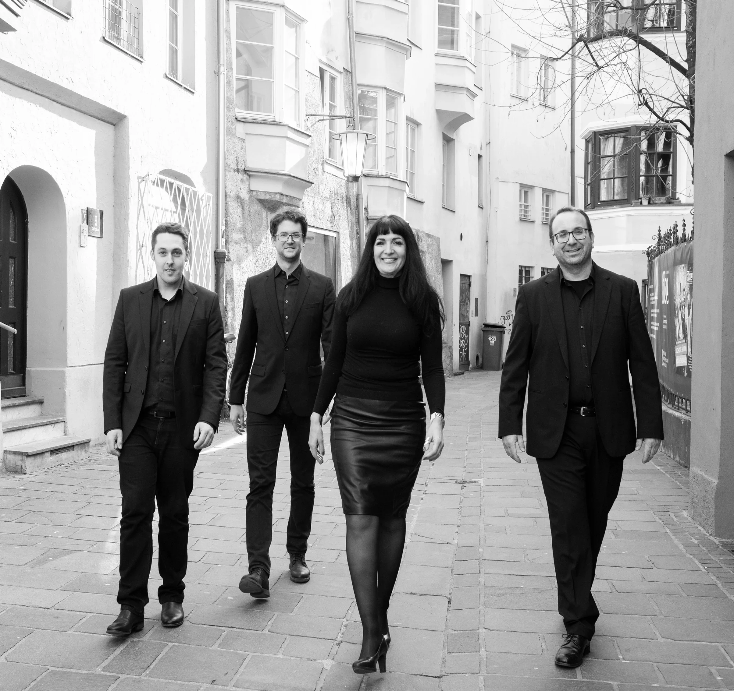 Gruppen von vier Menschen, drei Männer und eine Frau, gehen in Schwarz-Weiß-Bildung auf einer Kopfsteinpflasterstraße. Sie tragen formelle Kleidung und lachen.