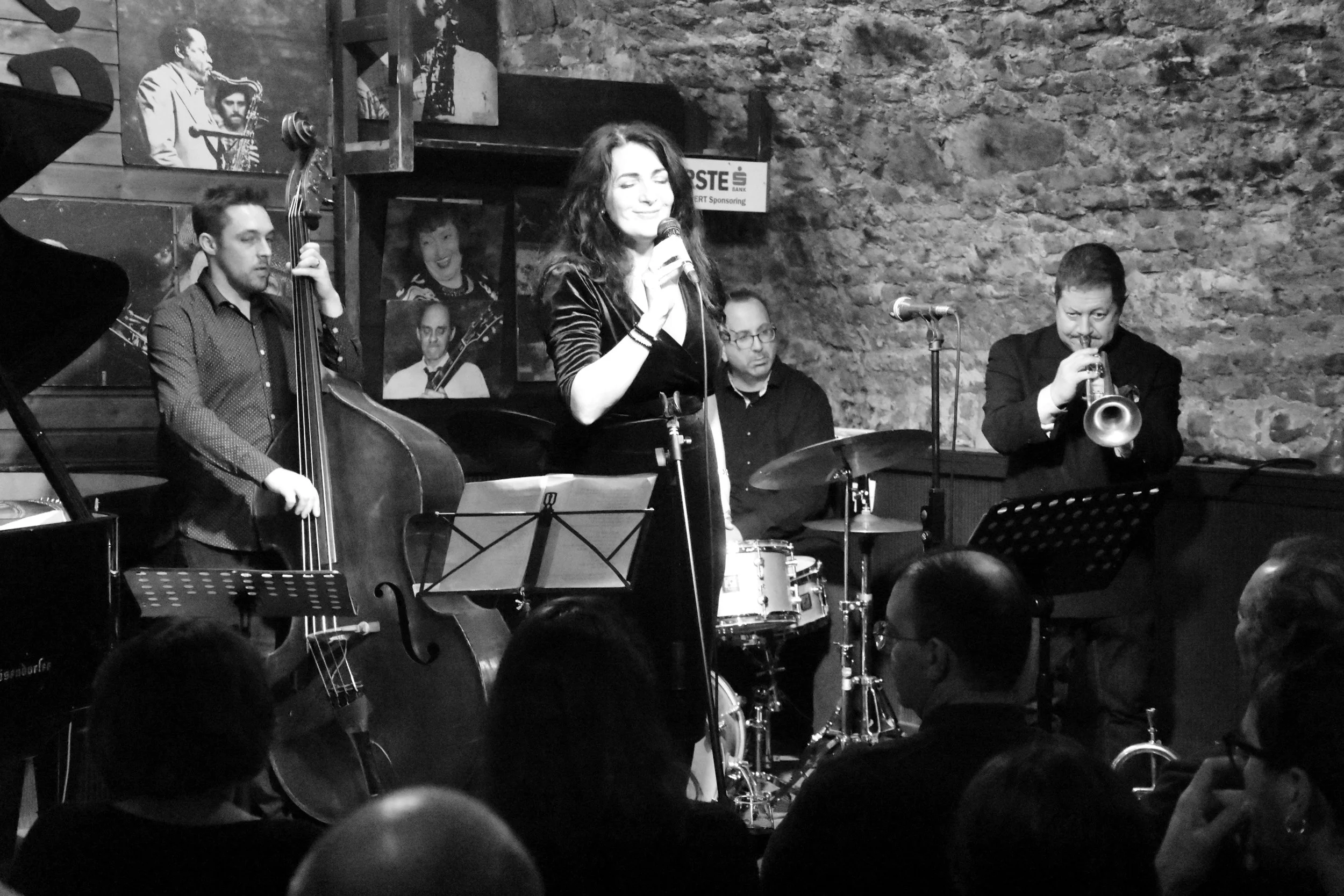 Eine Frau singt mit einem Mikrofon, begleitet von einer Jazzband mit Bass, Schlagzeug und Trompete, in einer Bar vor einer rauen Ziegelwand, im Schwarz-Weiß-Stil.