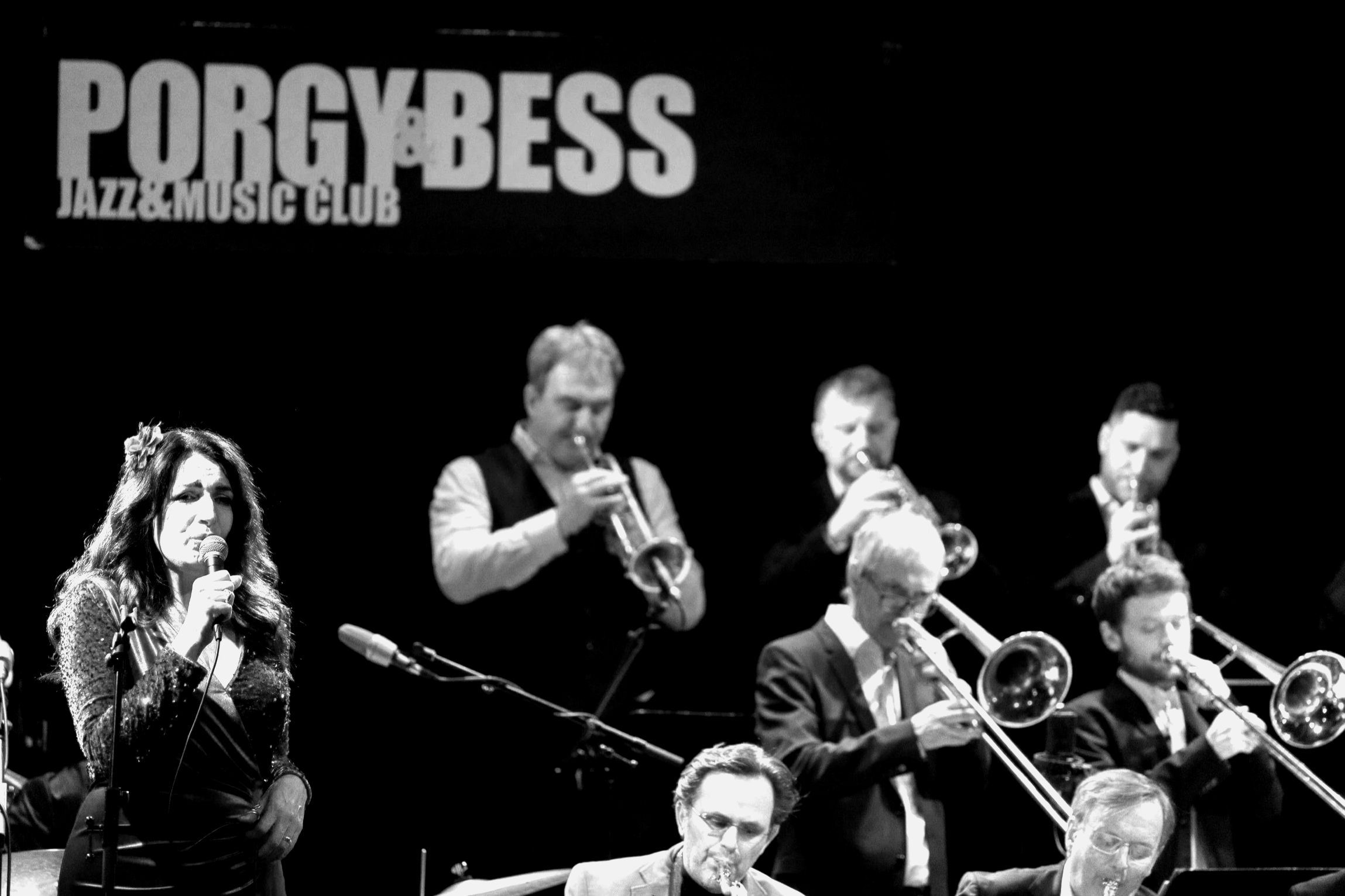 Junge Sängerin mit Mikrofon und ein kleines Jazzorchester mit Trompeten, Saxophon und Klavier auf einer Bühne bei Porgy&Bess, einem Jazz & Music Club.