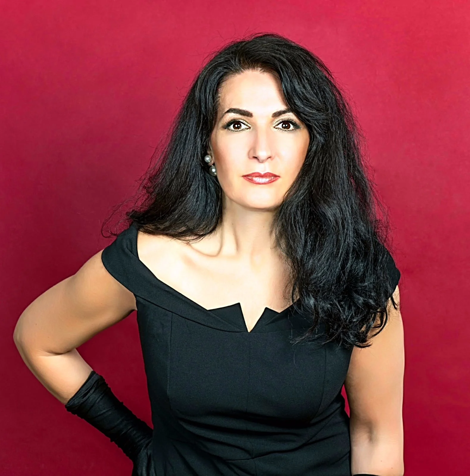 eine Frau mit schwarzen, lockigen Haaren und roten Lippen trägt ein schwarzes Kleid mit einem V-Ausschnitt vor einem roten Hintergrund