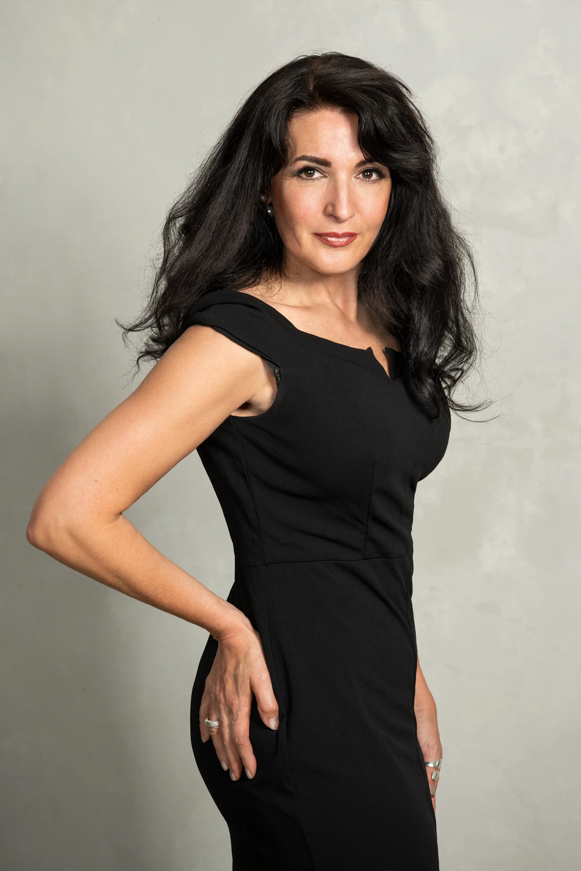Porträt einer Frau mit schwarzen, lockigen Haaren, die ein schwarzes Kleid trägt, vor neutralem Hintergrund.