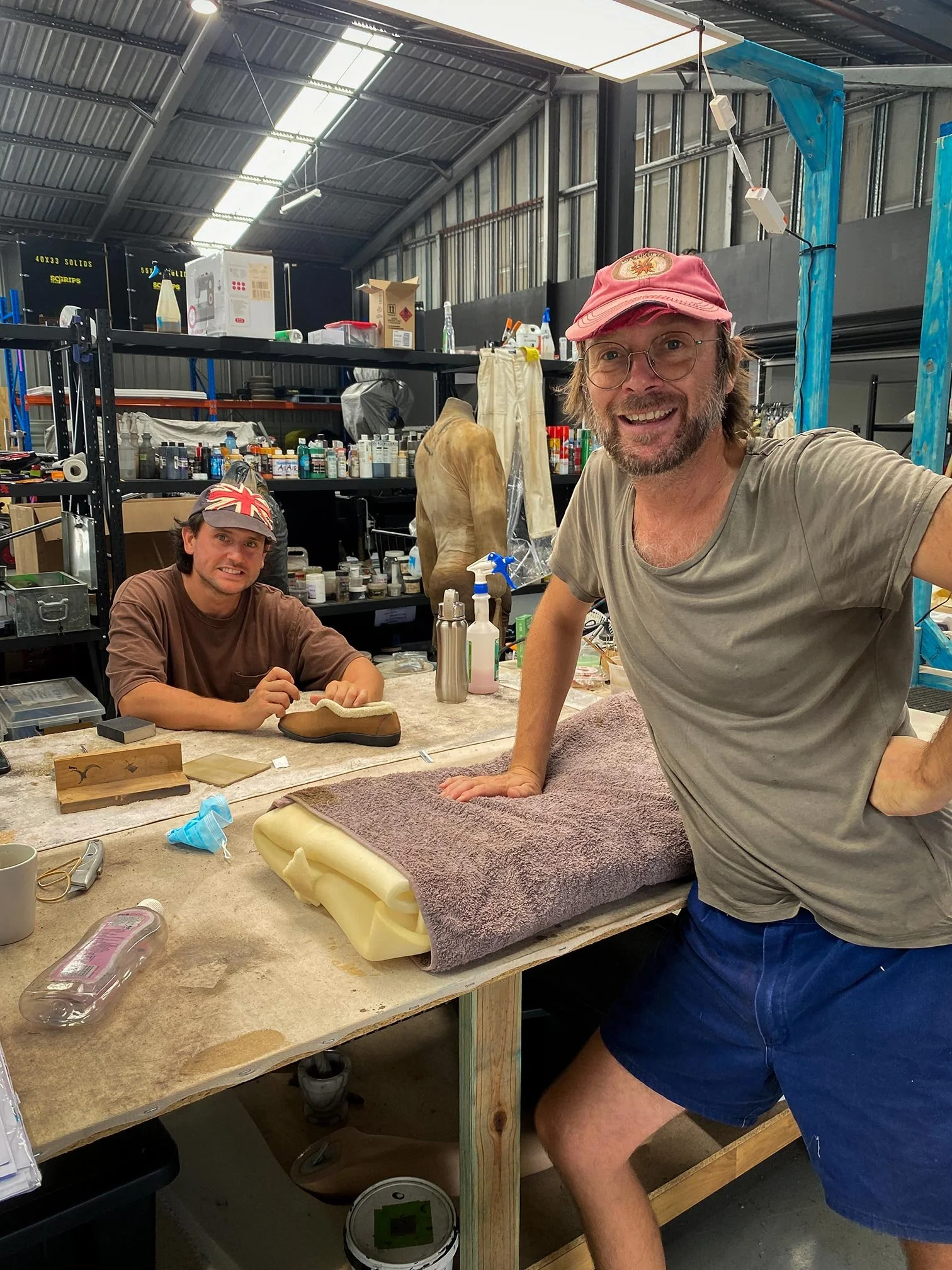 Two men in a workshop, one standing and smiling at the camera while leaning on a worktable, the other sitting and working on a shoe. The workshop has shelves with various supplies and tools in the background.