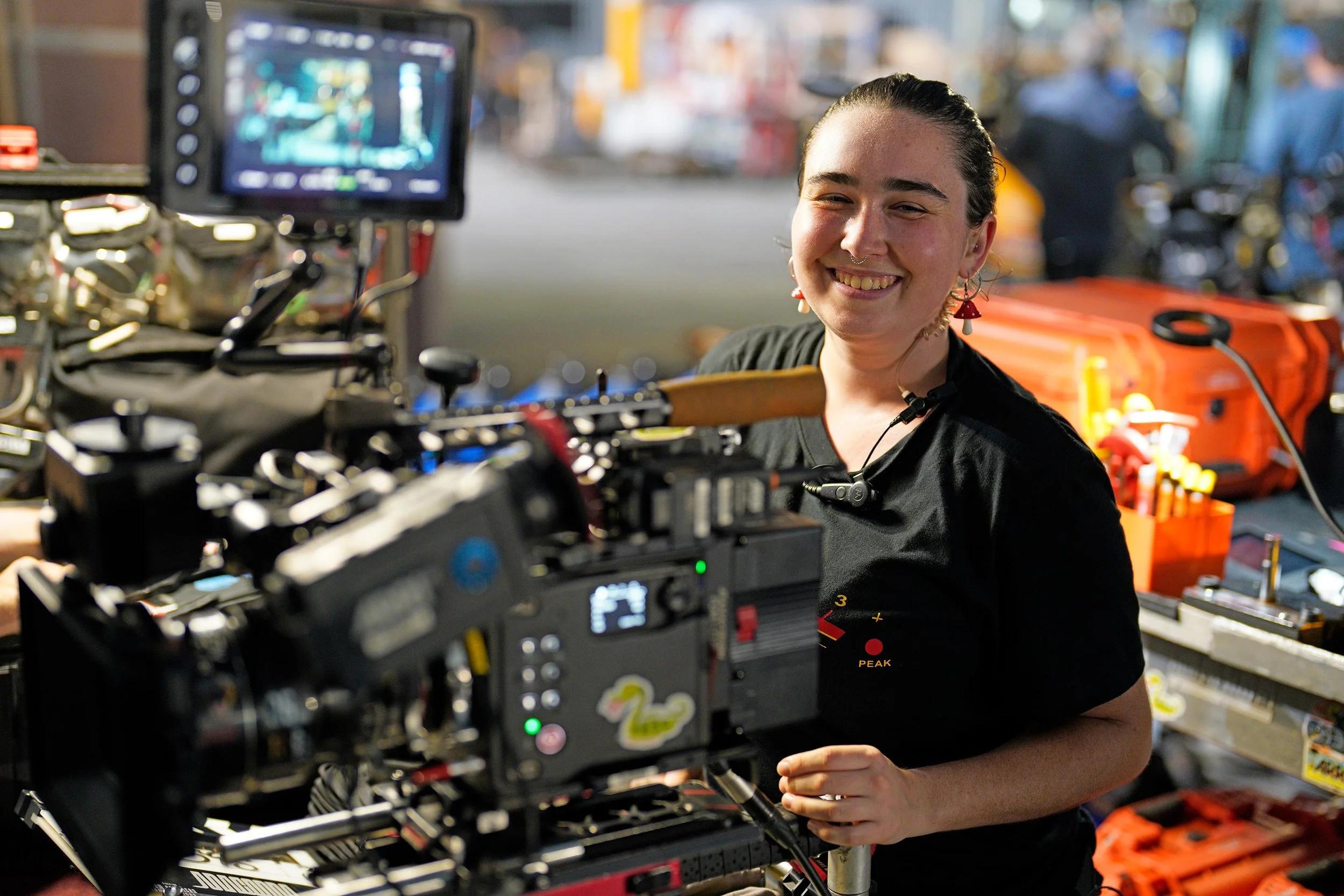 A woman with earrings and a piercing smiling behind a camera on a film set or production location with equipment and tools surrounding her.