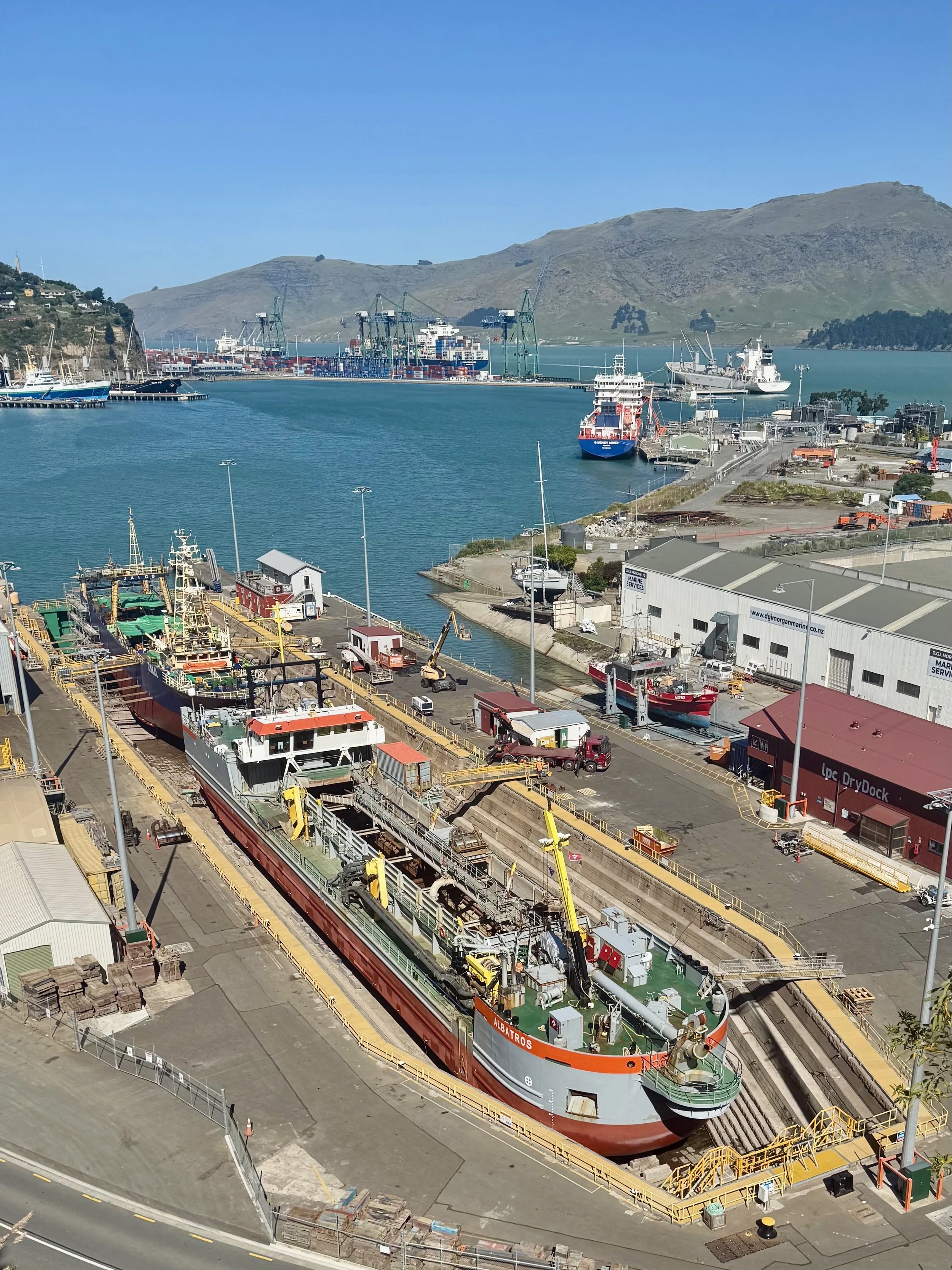 A harbor with ships docked, a large crane, hills in the background, and a ship being serviced in the foreground.