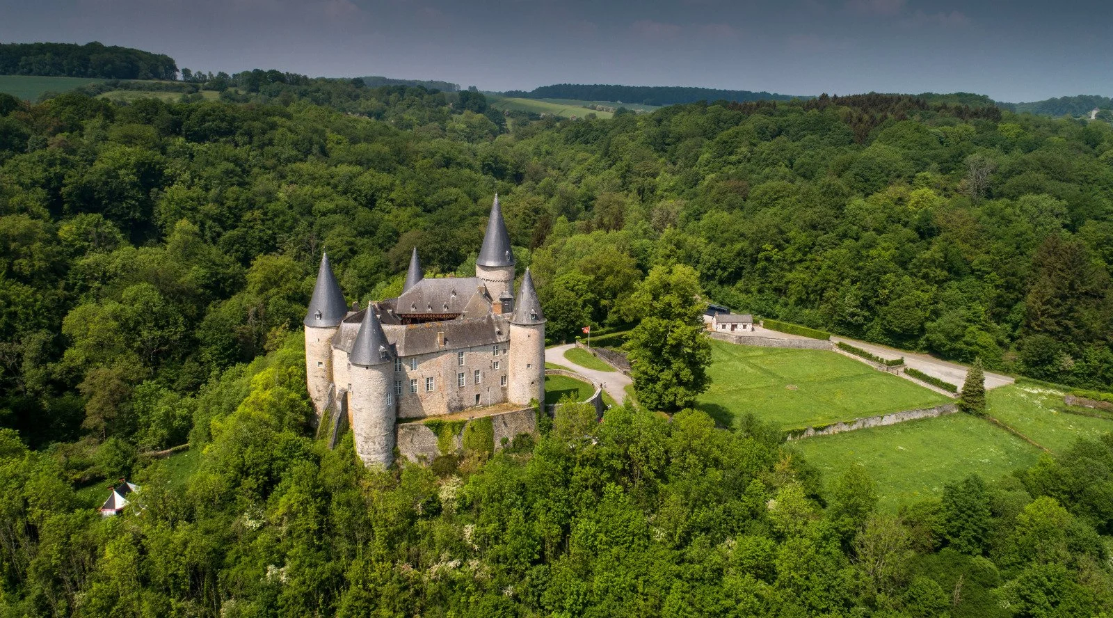 Un château médiéval entouré de forêt dense, avec des tours coniques en tuiles grises, situé sur une colline verdoyante. 