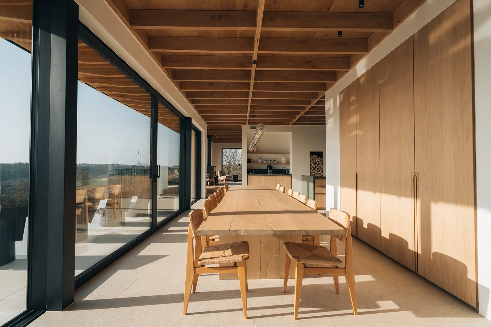 Salle à manger avec grande table en bois et chaises, près de grandes fenêtres coulissantes offrant une vue extérieure, intérieur moderne en bois clair et design épuré.