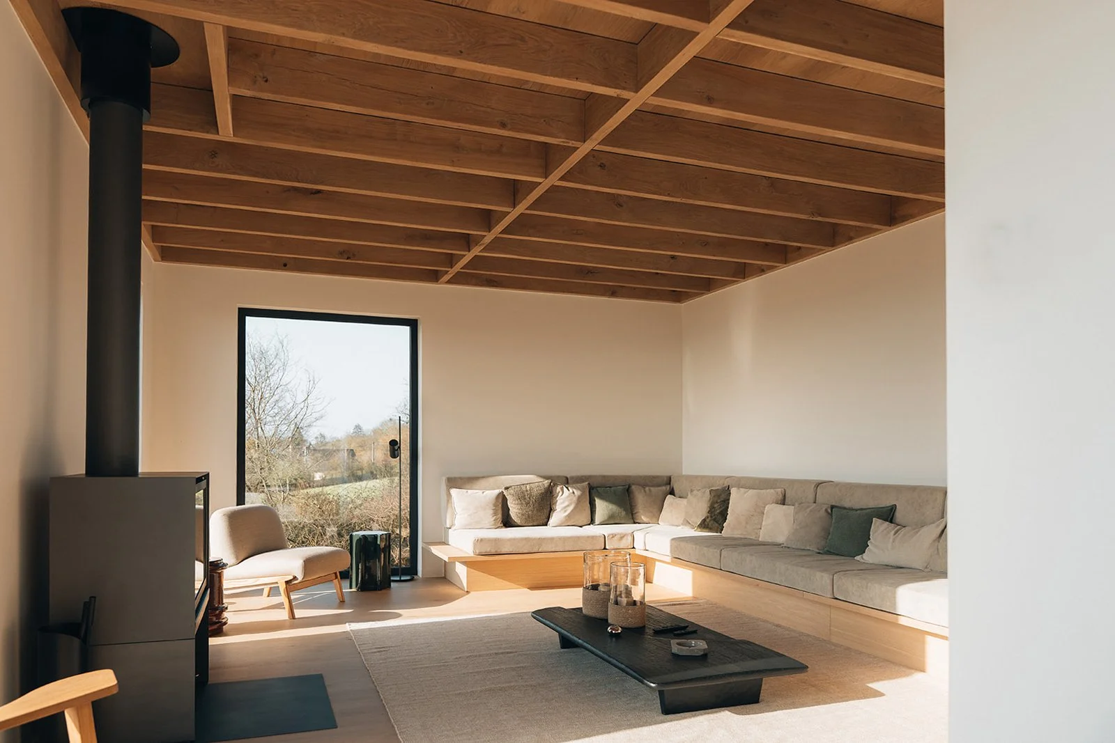 Salon moderne avec canapé beige, contre un mur blanc, sous un plafond en bois, avec une fenêtre donnant sur un jardin extérieur, table basse noire avec bougies, fauteuil beige, tapis beige