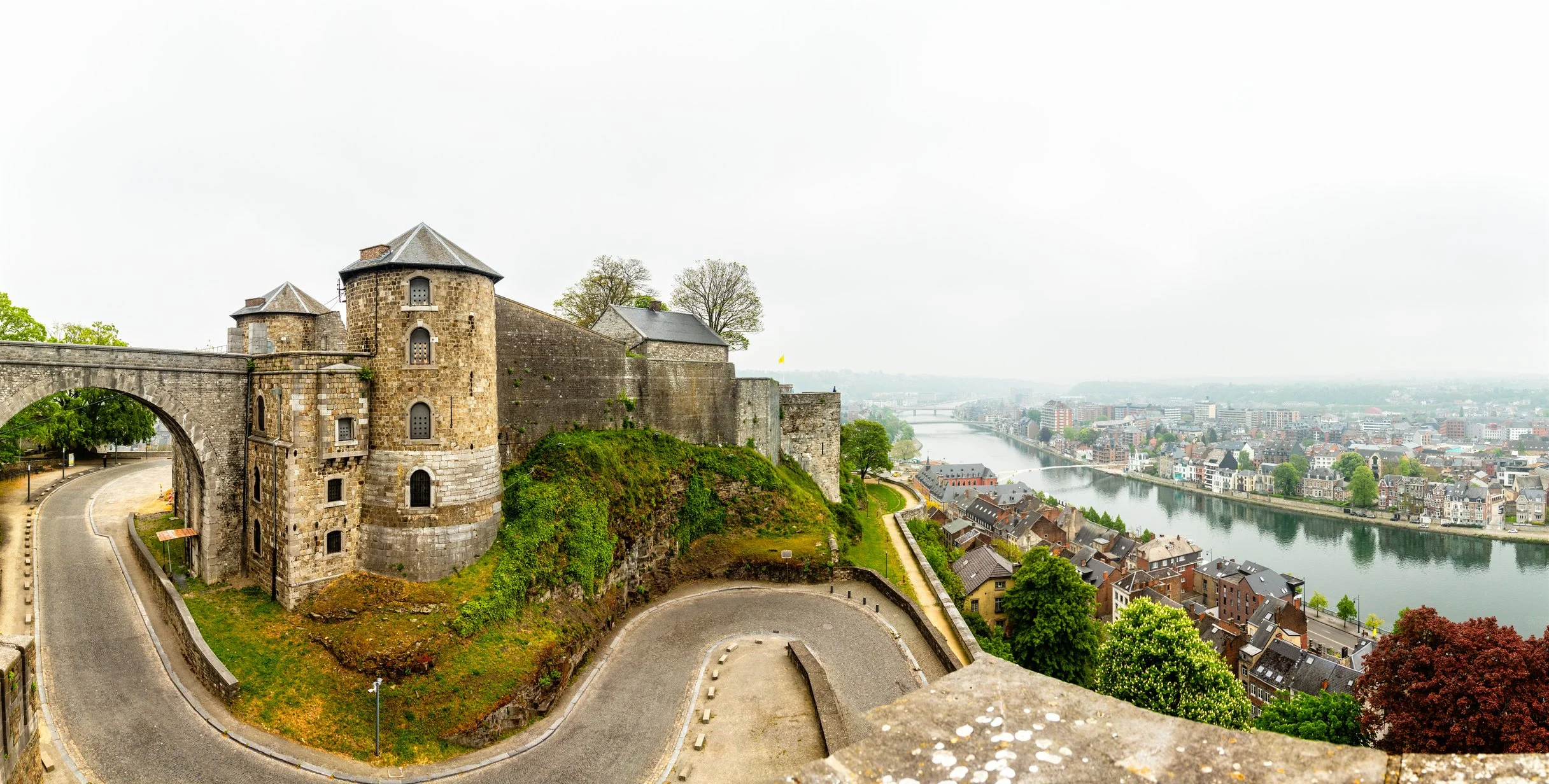 Vue panoramique d'un château médiéval en pierre au sommet d'une colline surplombant une ville et une rivière, avec des bâtiments modernes et des arbres verts dans un paysage urbain.