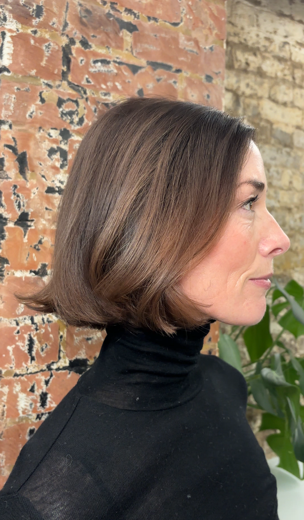 Side profile of a woman with shoulder-length straight brown hair, wearing a black turtleneck, standing against a rustic brick wall with some greenery.