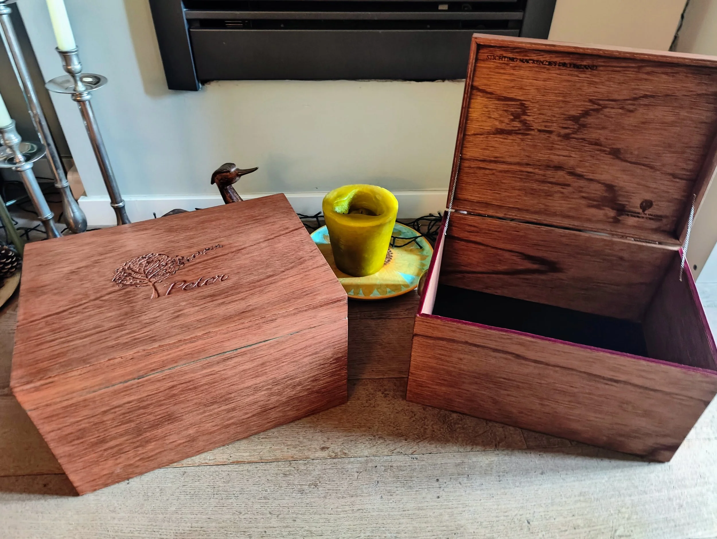 A wooden box with the name 'Peter' and a musical tree logo engraved on the lid. There is an open wooden box beside it, showing its interior. Both are mahogany patina stained and waxed to enhance the intricate Okoume wood grain structure. .