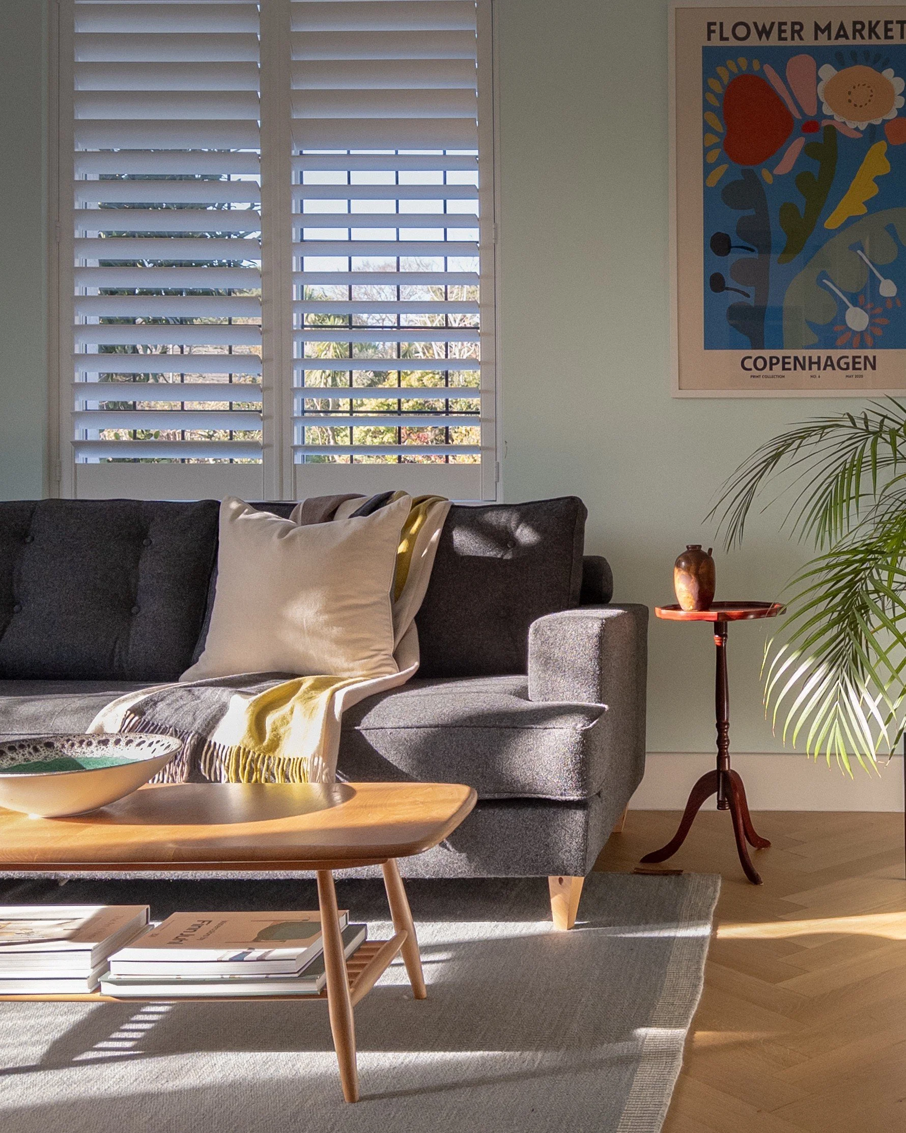 Living room with a gray sofa adorned with pillows and a blanket, a wooden coffee table with magazines, a side table with a small vase, a window with white shutters, a potted plant, and a colorful flower market poster on the wall.
