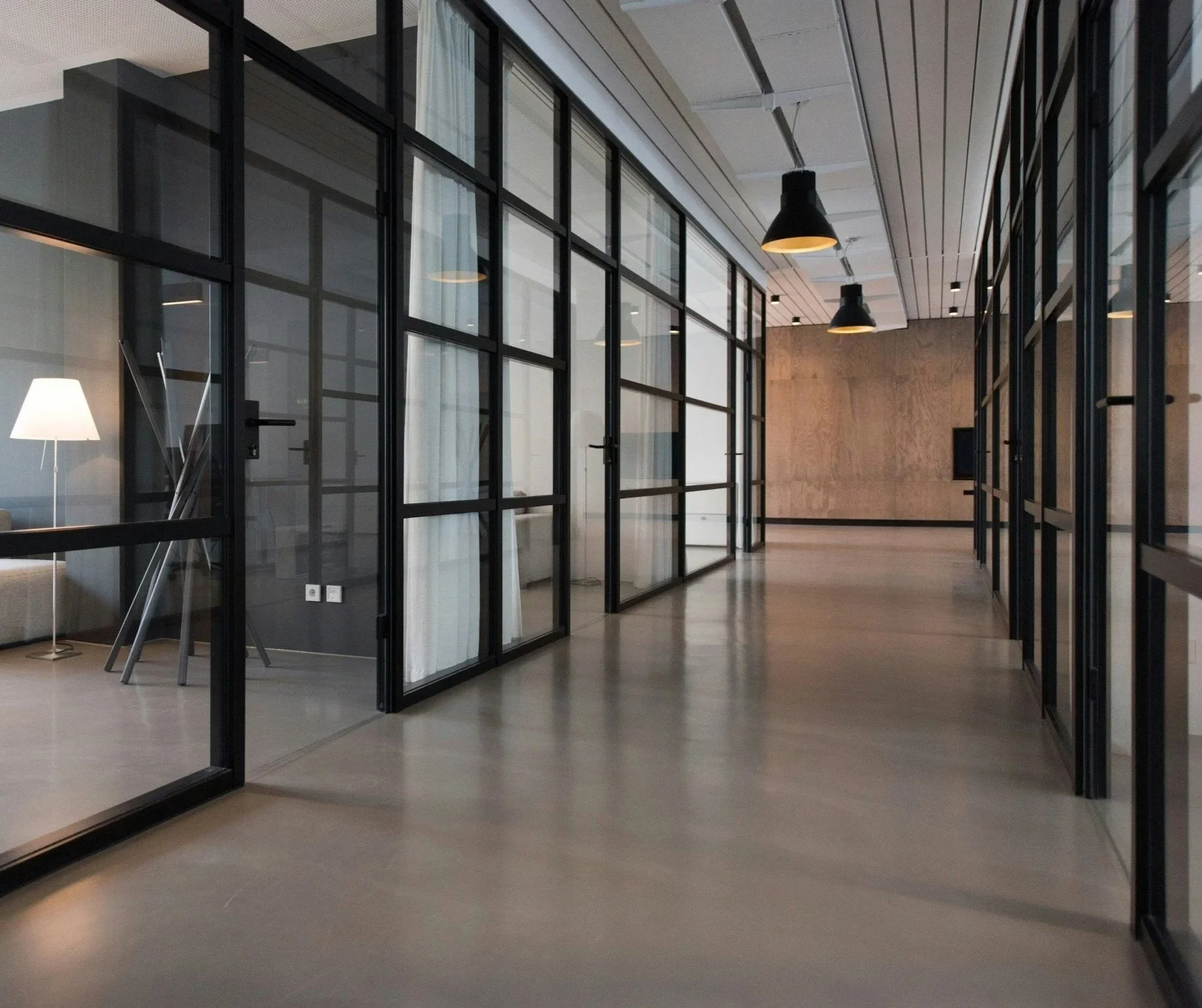 Modern office corridor with glass walls and black framing, featuring black pendant lights and a wooden accent wall at the end.