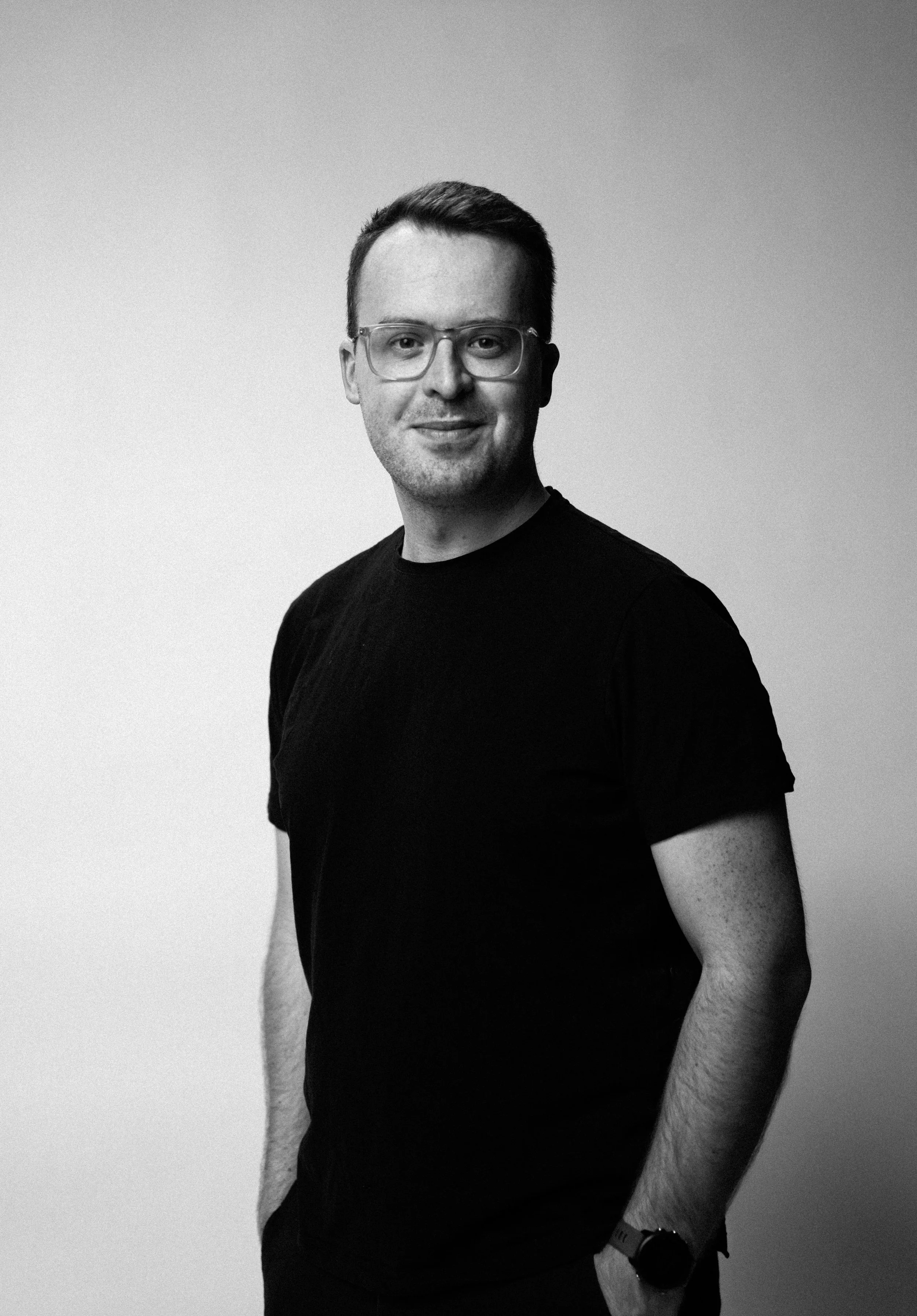 Schwarz-Weiß-Foto eines lächelnden jungen Mannes mit Brille, schwarzer T-Shirt, steht vor hellem, neutralem Hintergrund.