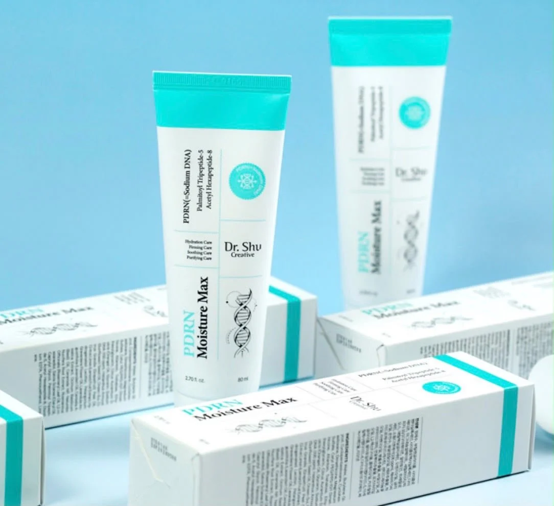 Multiple white tubes of skincare product with turquoise caps and accents, labeled 'PDRN Moisture Max' and 'Dr. Shu Creative,' alongside their white boxes on a light blue background.